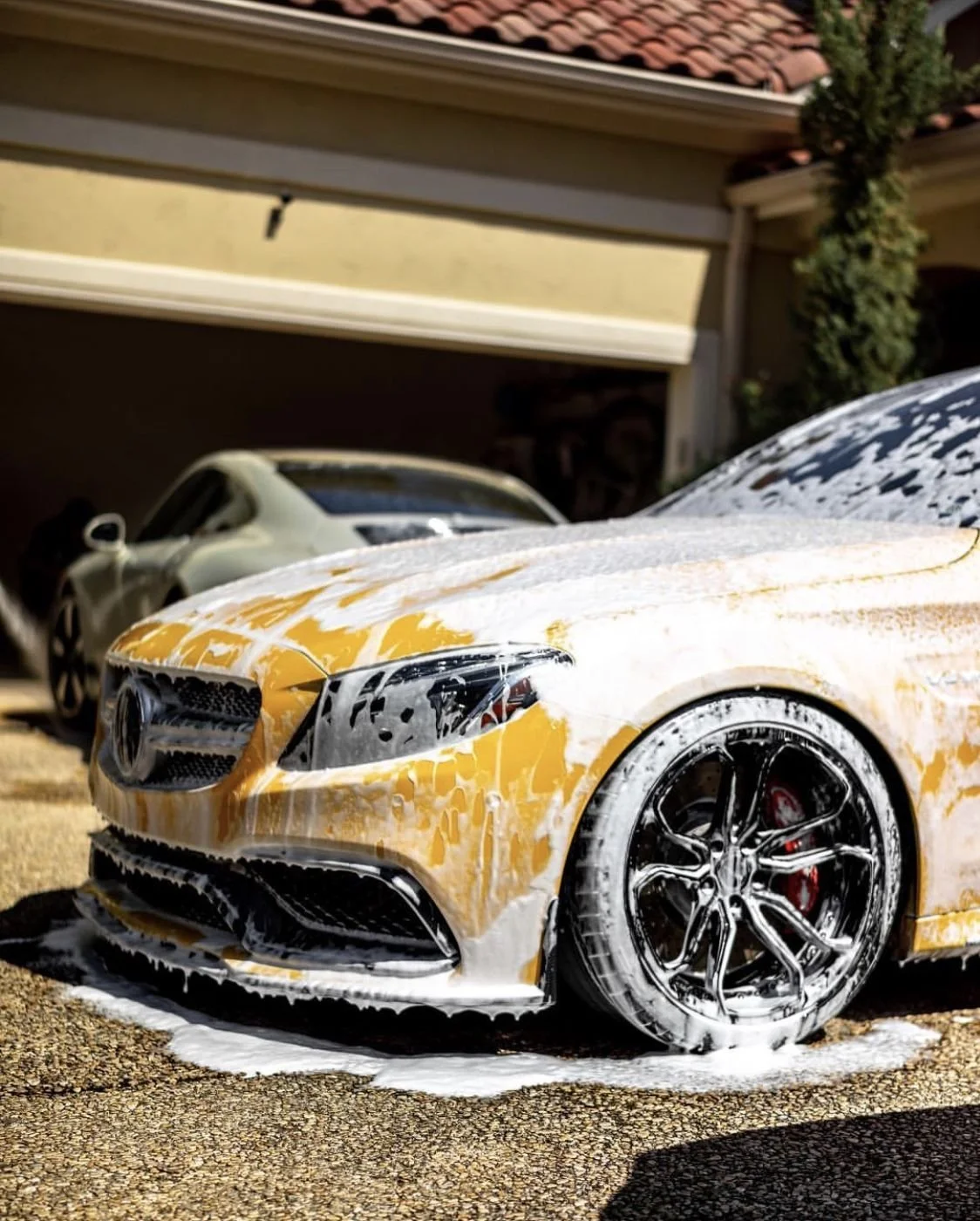Yellow sports car covered in soap and foam during a car wash, with a black car in the background in a driveway.