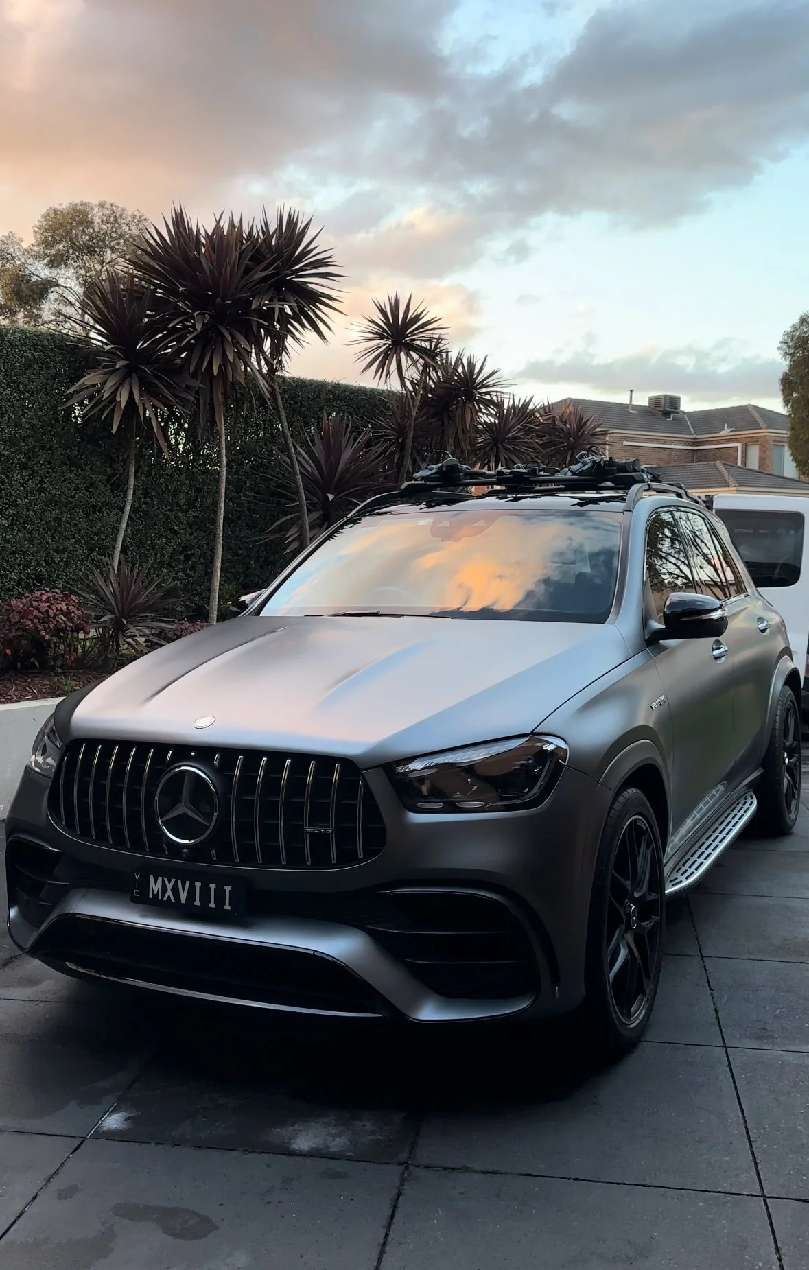 A silver Mercedes-Benz SUV parked on a driveway during sunset, with palm-like trees and a house in the background.