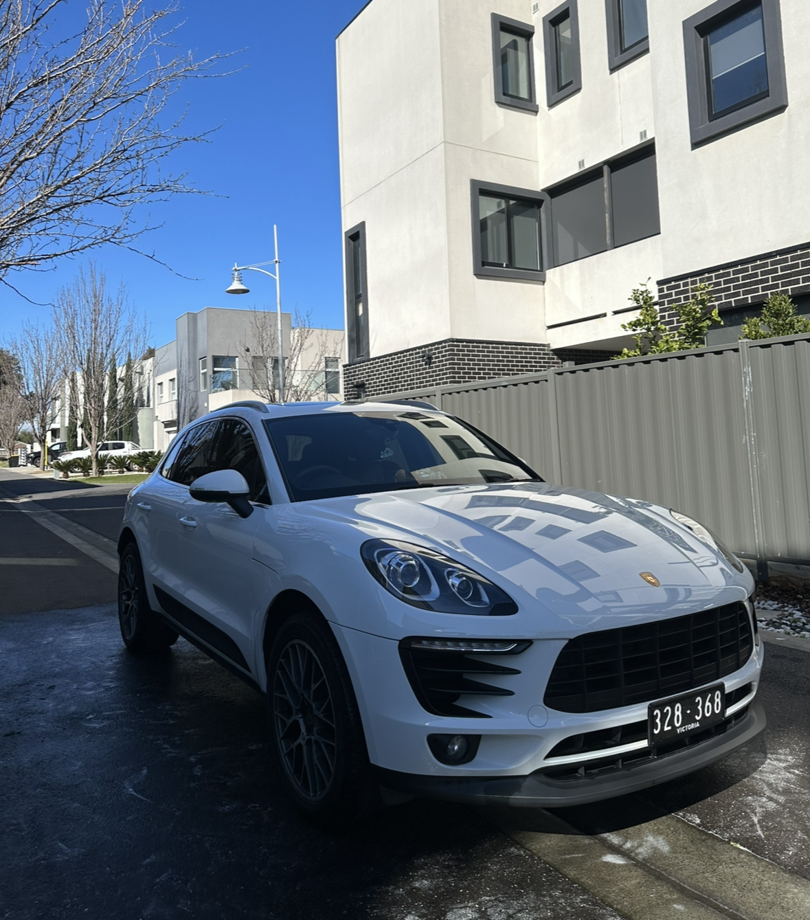 White Porsche SUV parked on a street in front of contemporary residential buildings on a clear, sunny day.