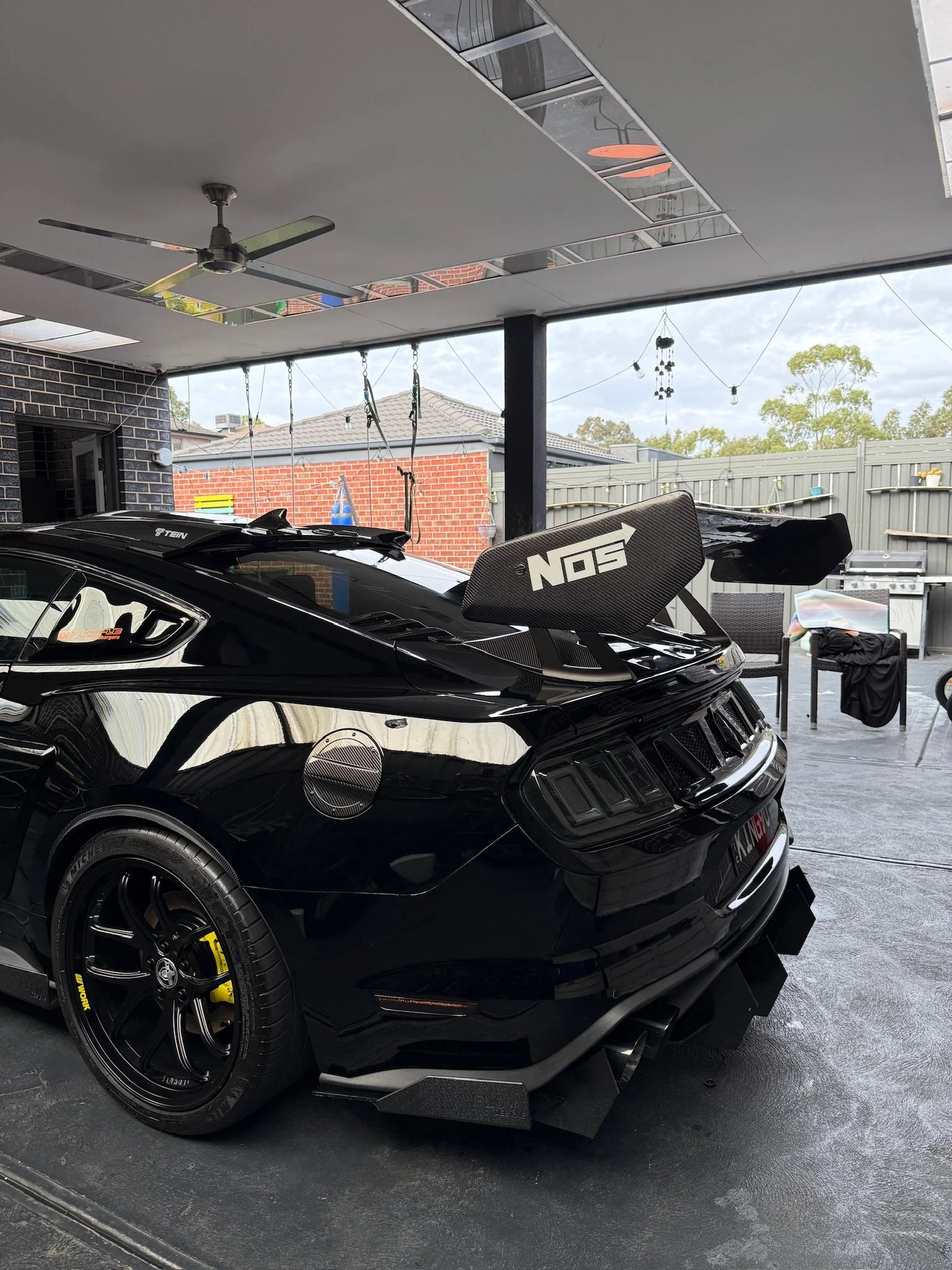Black race car with a large rear wing and racing tires parked on a driveway under a covered area. The car is in front of a brick wall and a fence, with outdoor decorations and a grill visible in the background.