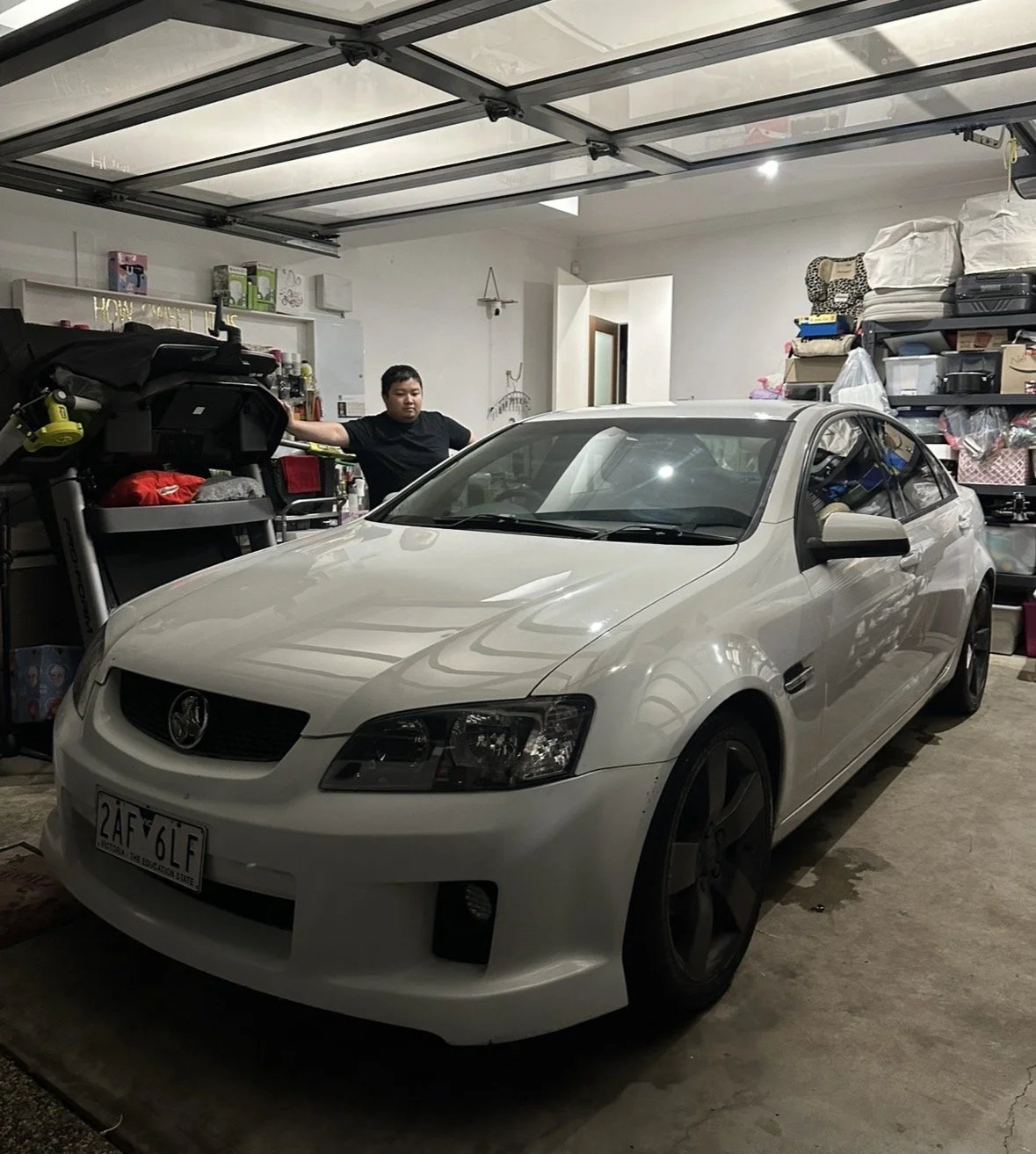 A white car parked inside a cluttered garage with a person standing behind it, surrounded by various items and storage shelves.