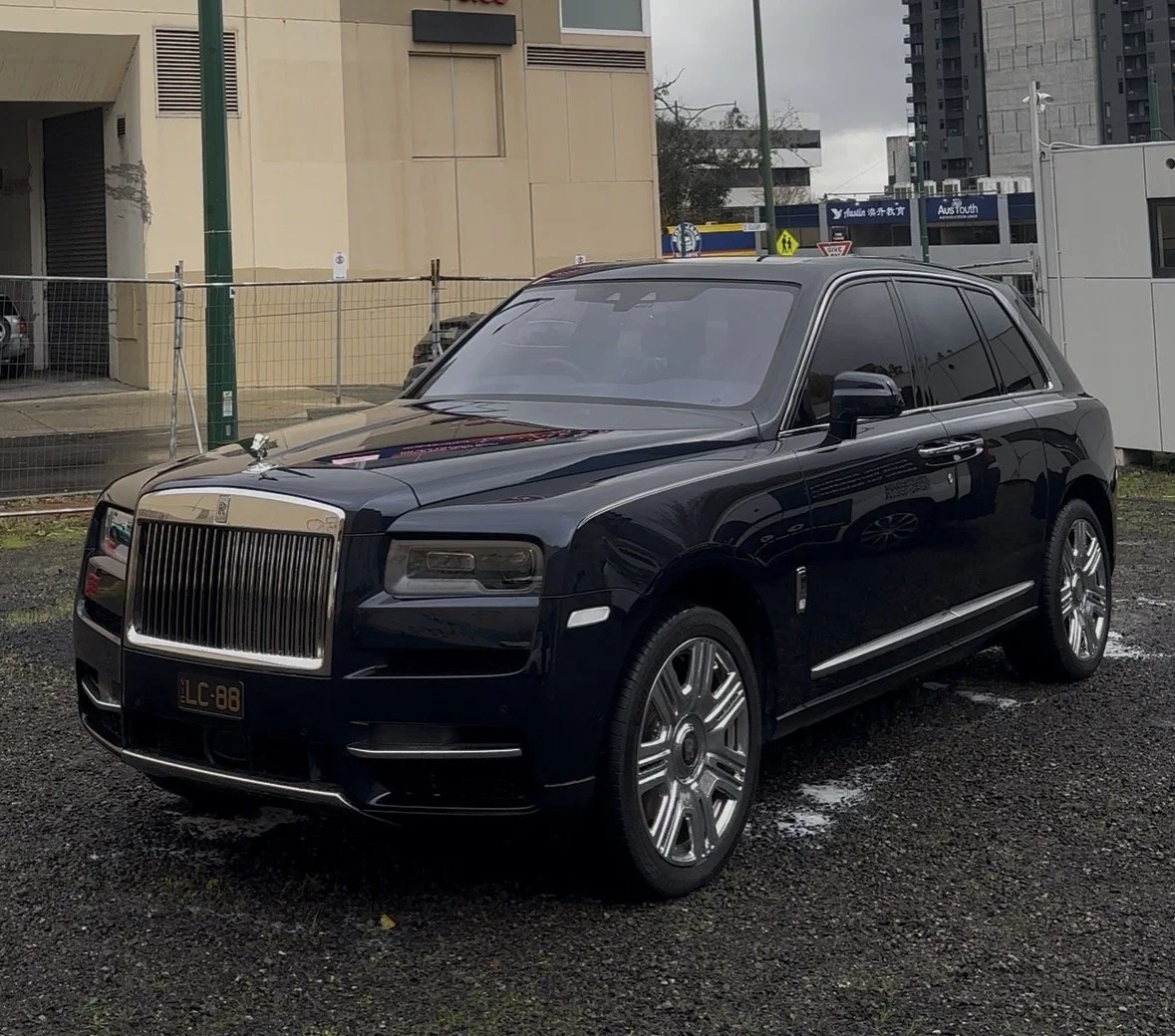 A black luxury SUV, specifically a Rolls-Royce Cullinan, parked on an outdoor gravel lot with buildings and cityscape in the background.