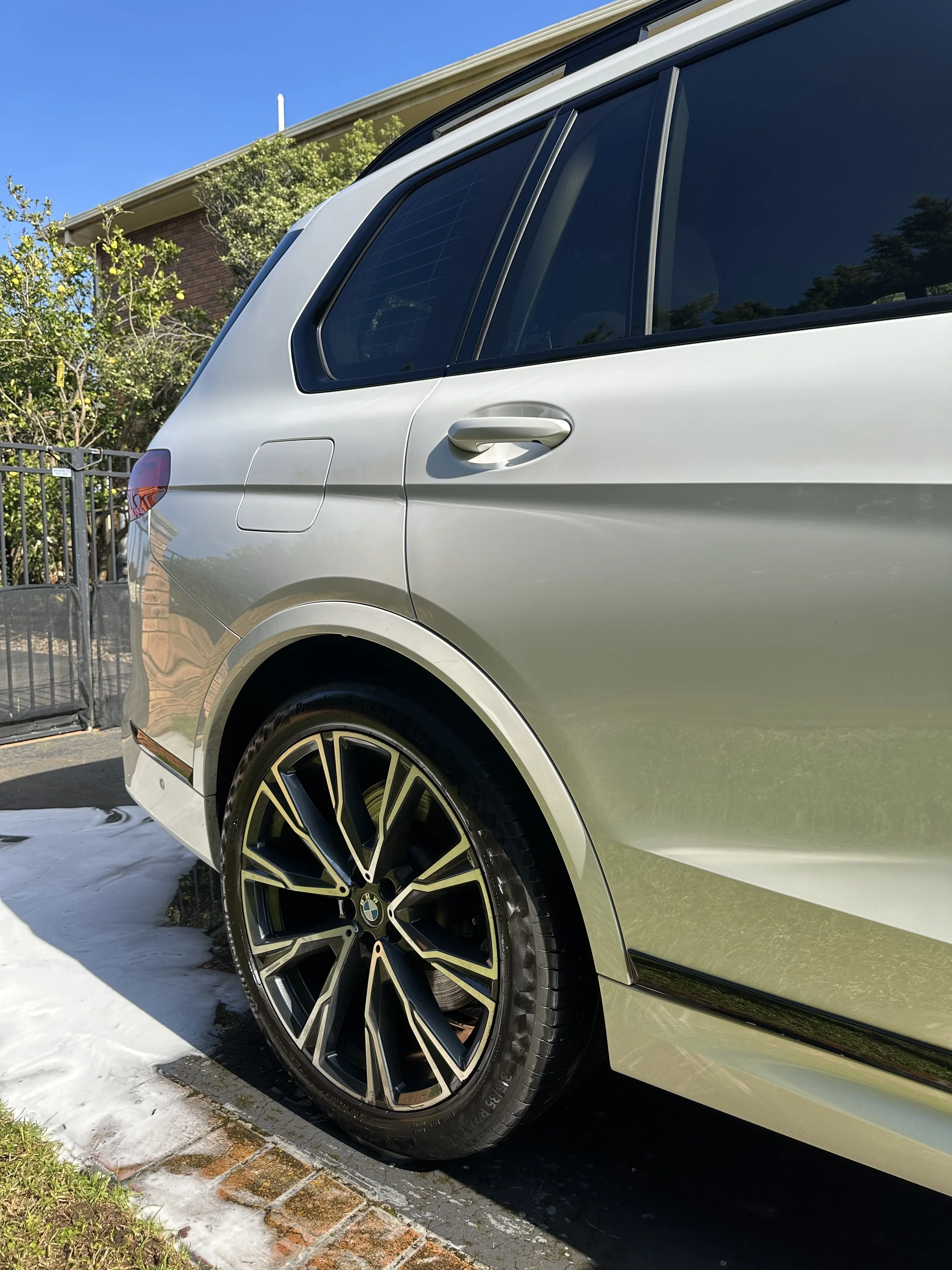 Close-up of the rear side of a silver BMW SUV parked on a driveway, with a black and gold rim wheel, a brick house, some greenery, and a blue sky in the background.