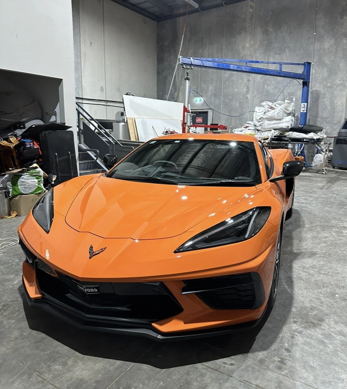 Orange Chevrolet Corvette sports car parked in an industrial-style garage with bags, boxes, and equipment in the background.