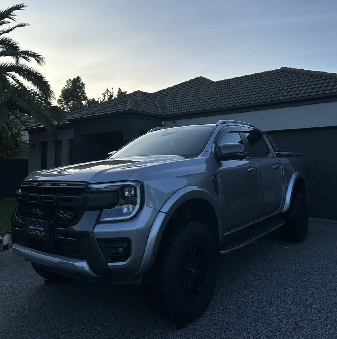 A silver pickup truck parked in front of a modern house with a dark roof and garage doors, with palm trees in the background during dusk.