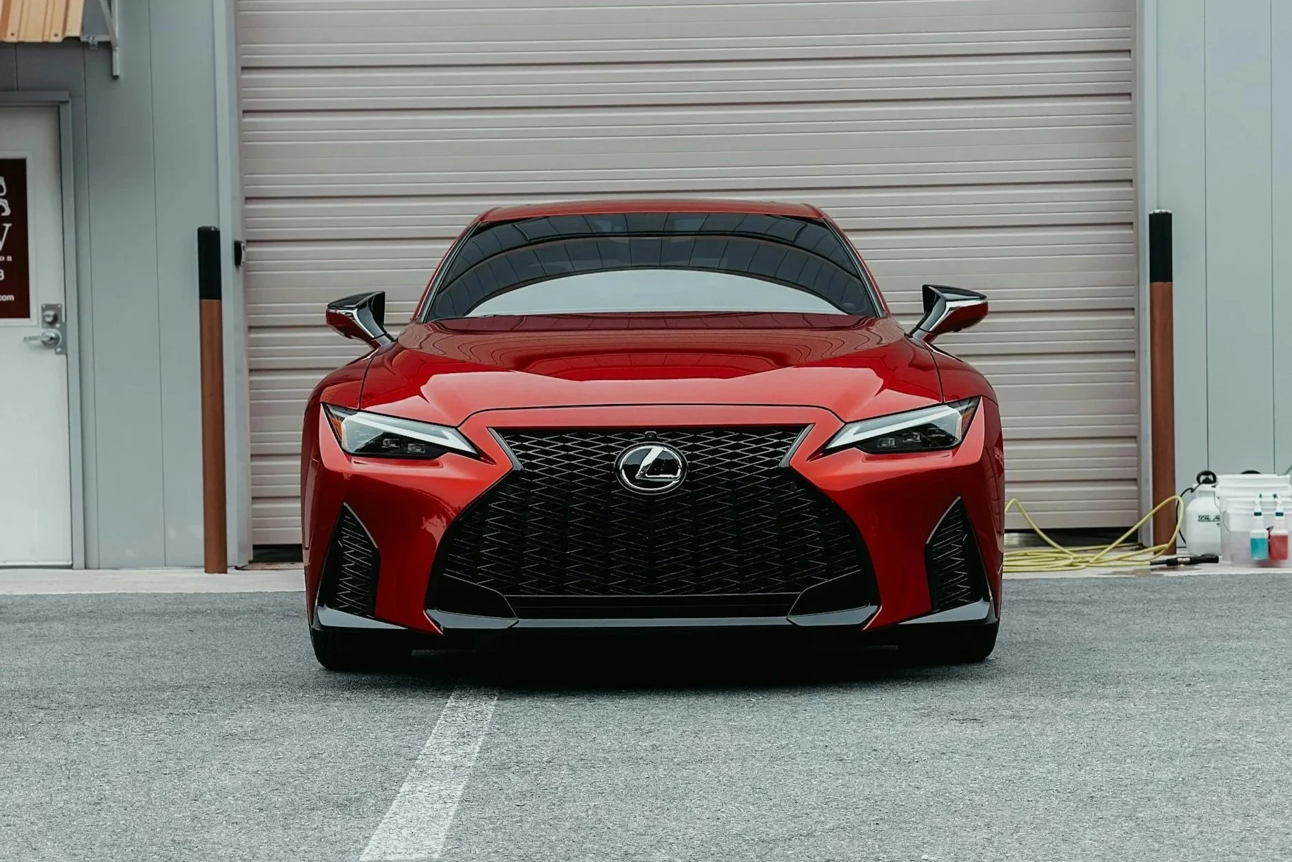 Red Lexus sports car parked in front of a beige garage with a roll-up door, adjacent to a garage door with a sign, and a white door on the side.
