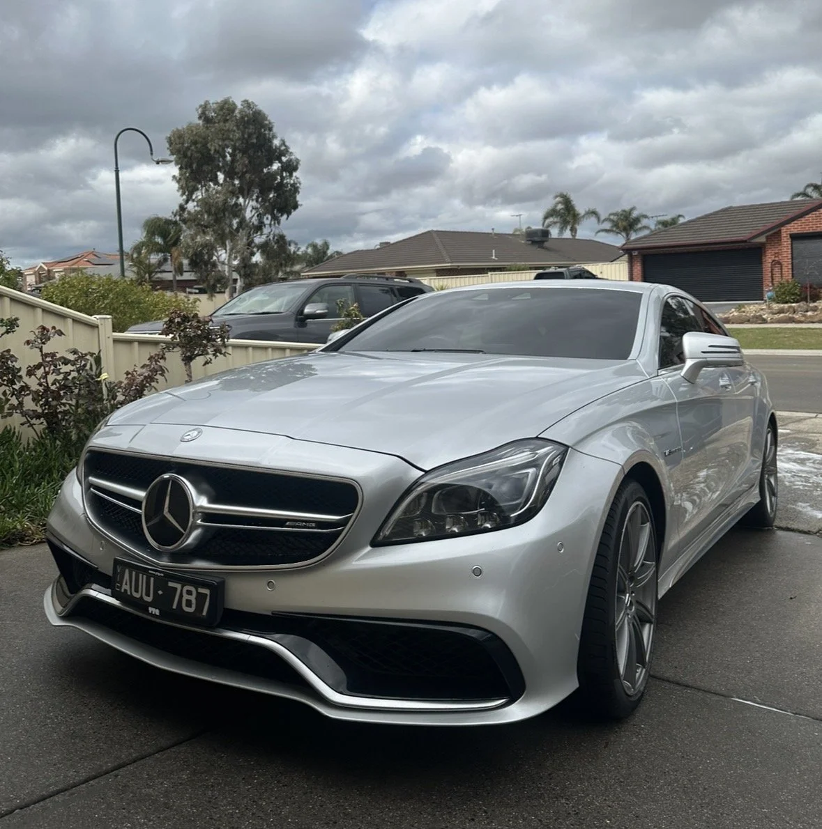 Silver Mercedes-Benz coupe parked on driveway in a suburban neighborhood, cloudy sky overhead, houses, trees, and a street in the background.