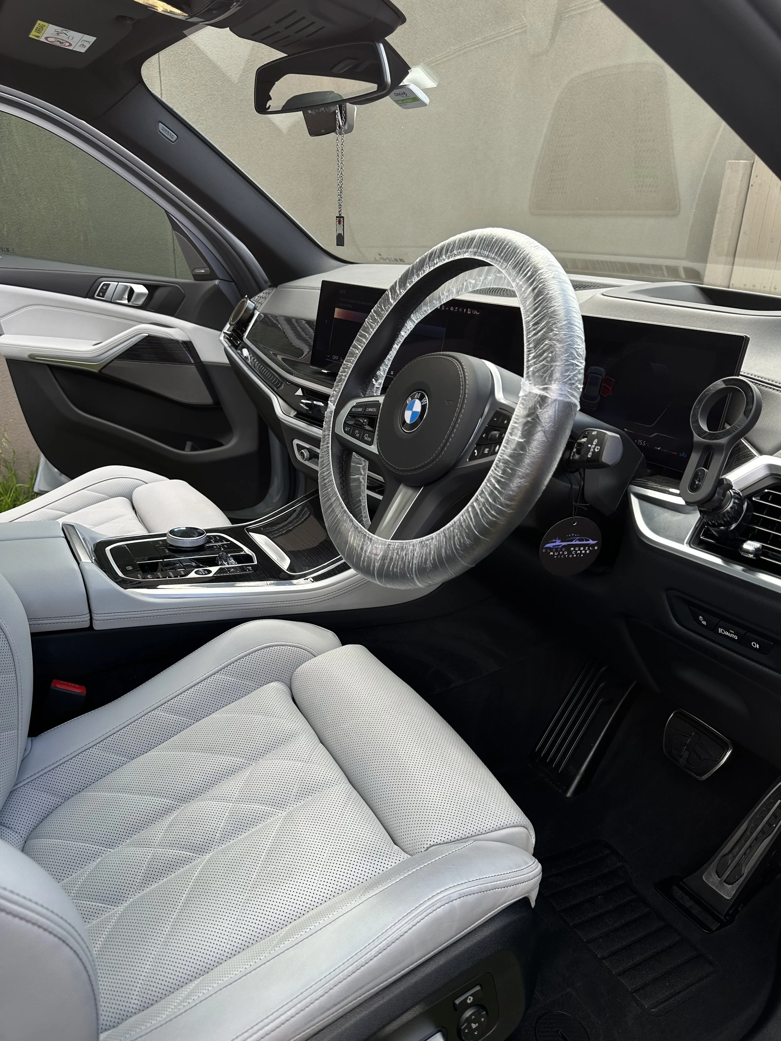The interior of a car showing a steering wheel with a plastic cover, a digital dashboard, a center console with controls, white leather seats, and a mirror with hanging ornaments.