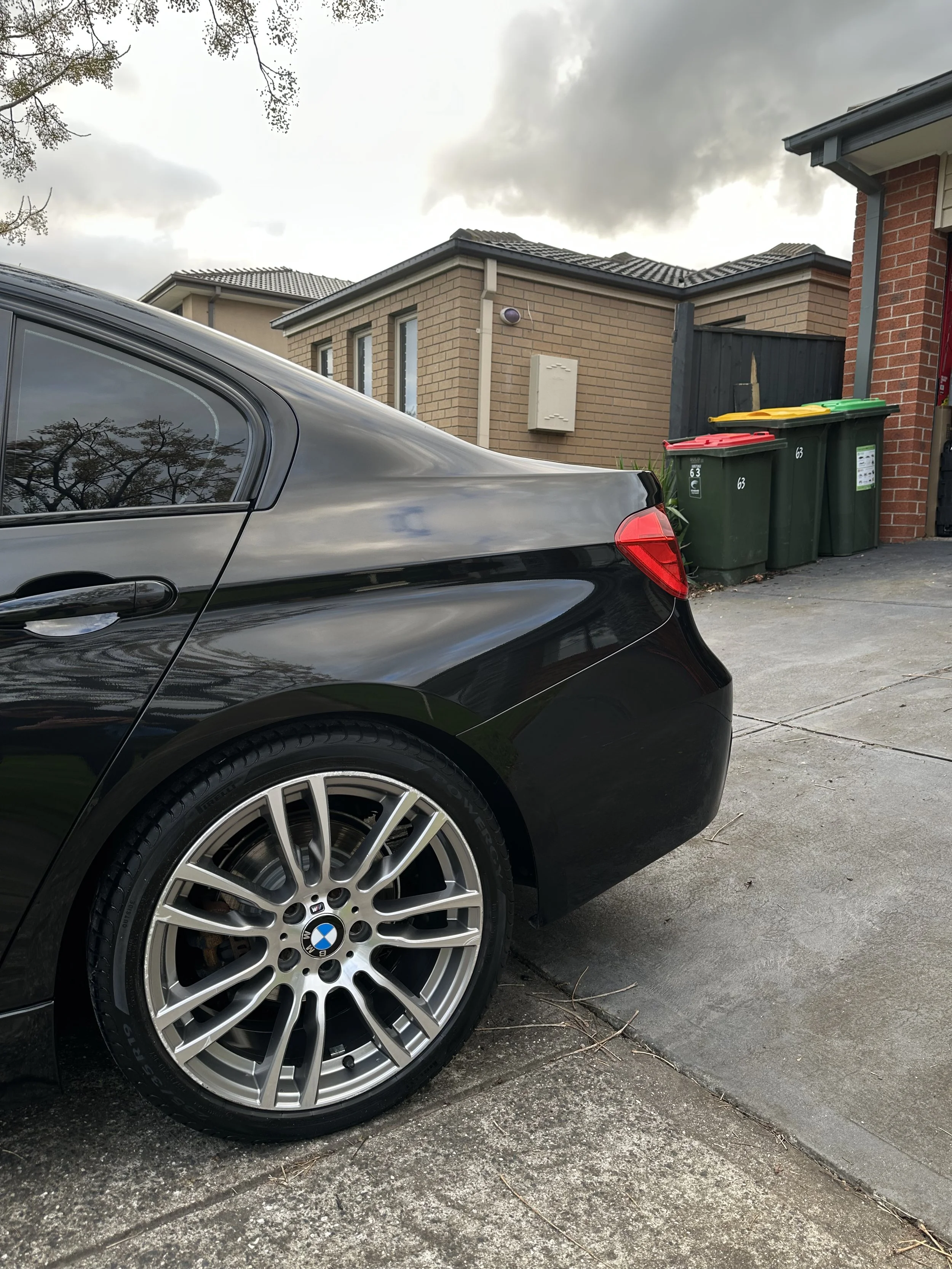 A close-up of the rear side of a black BMW car parked in a driveway next to a house. The car is shiny and has a detailed wheel rim. In the background, there are green trash bins with yellow, red, and green lids. Part of the house's brick exterior, windows, and roof with dark tiles are visible. The sky overhead is cloudy.