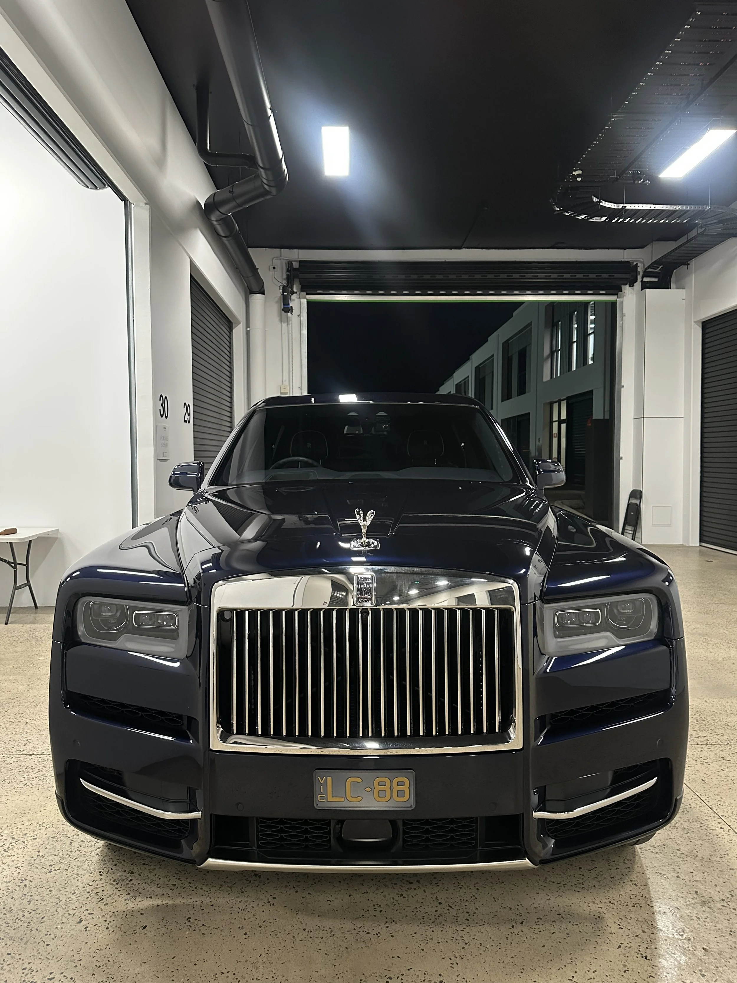 Black luxury car with a chrome grille, parked indoors in a garage, with a black hood ornament on the front.
