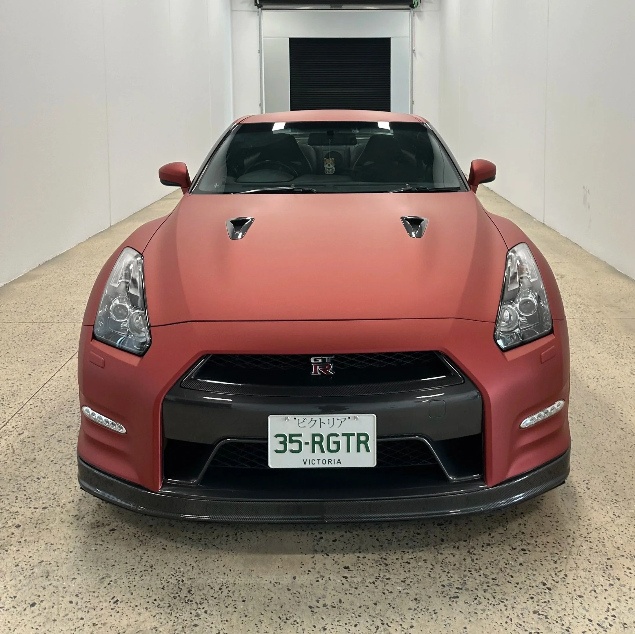Front view of a red Nissan GT-R sports car parked inside a garage with white walls and speckled beige floor. The car has a Japanese license plate from Victoria, Australia.