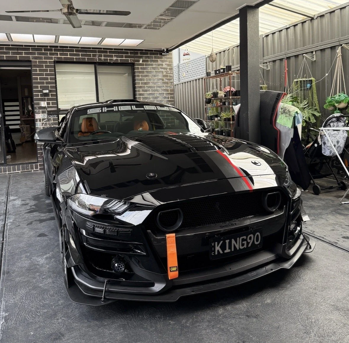 Black Shelby sports car with orange accents, parked in a garage patio, with various plants and outdoor furniture on the side.