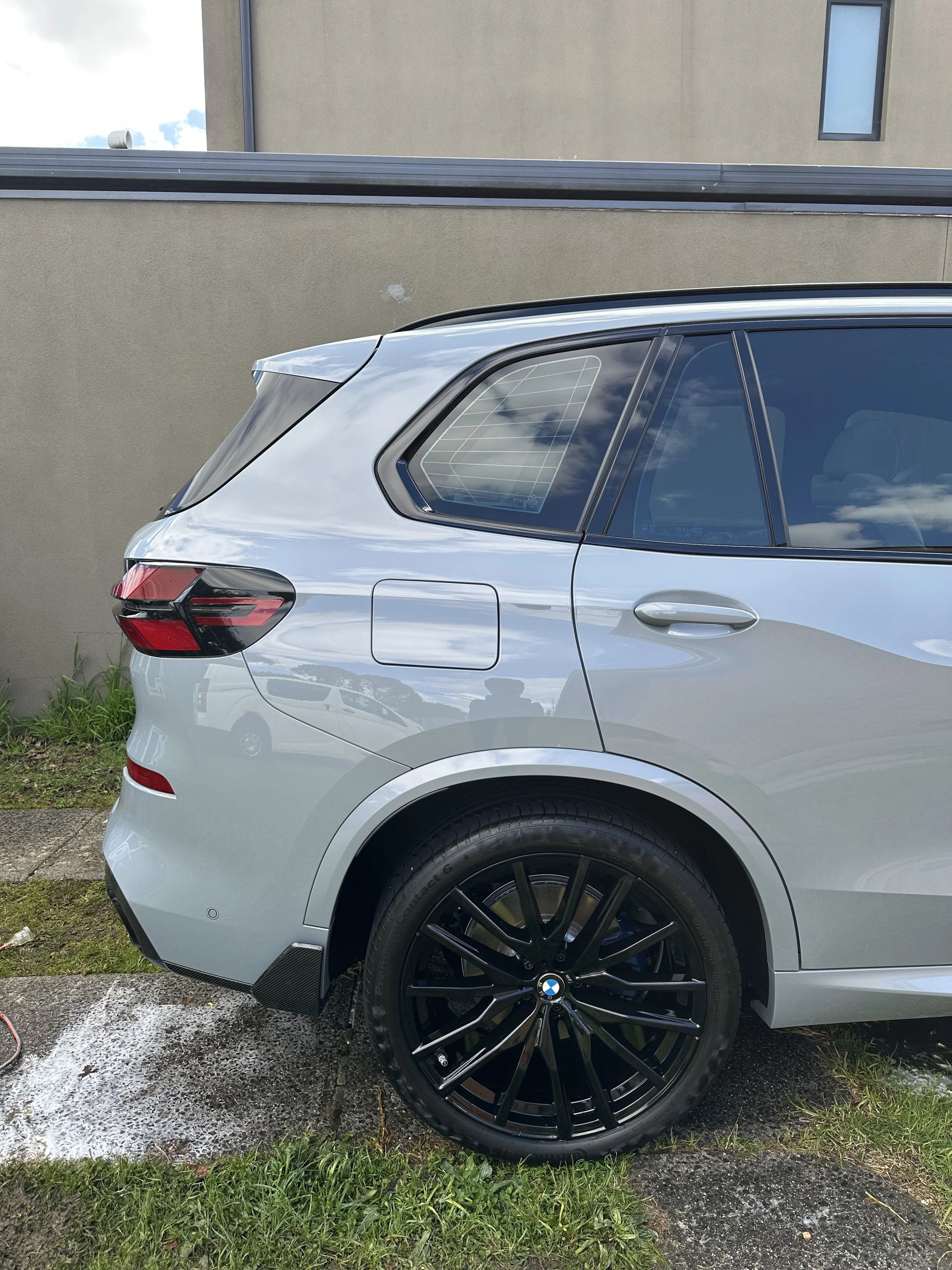 Close-up side view of a silver BMW car parked on wet pavement with black alloy wheels, against a beige building wall and grass in the foreground.