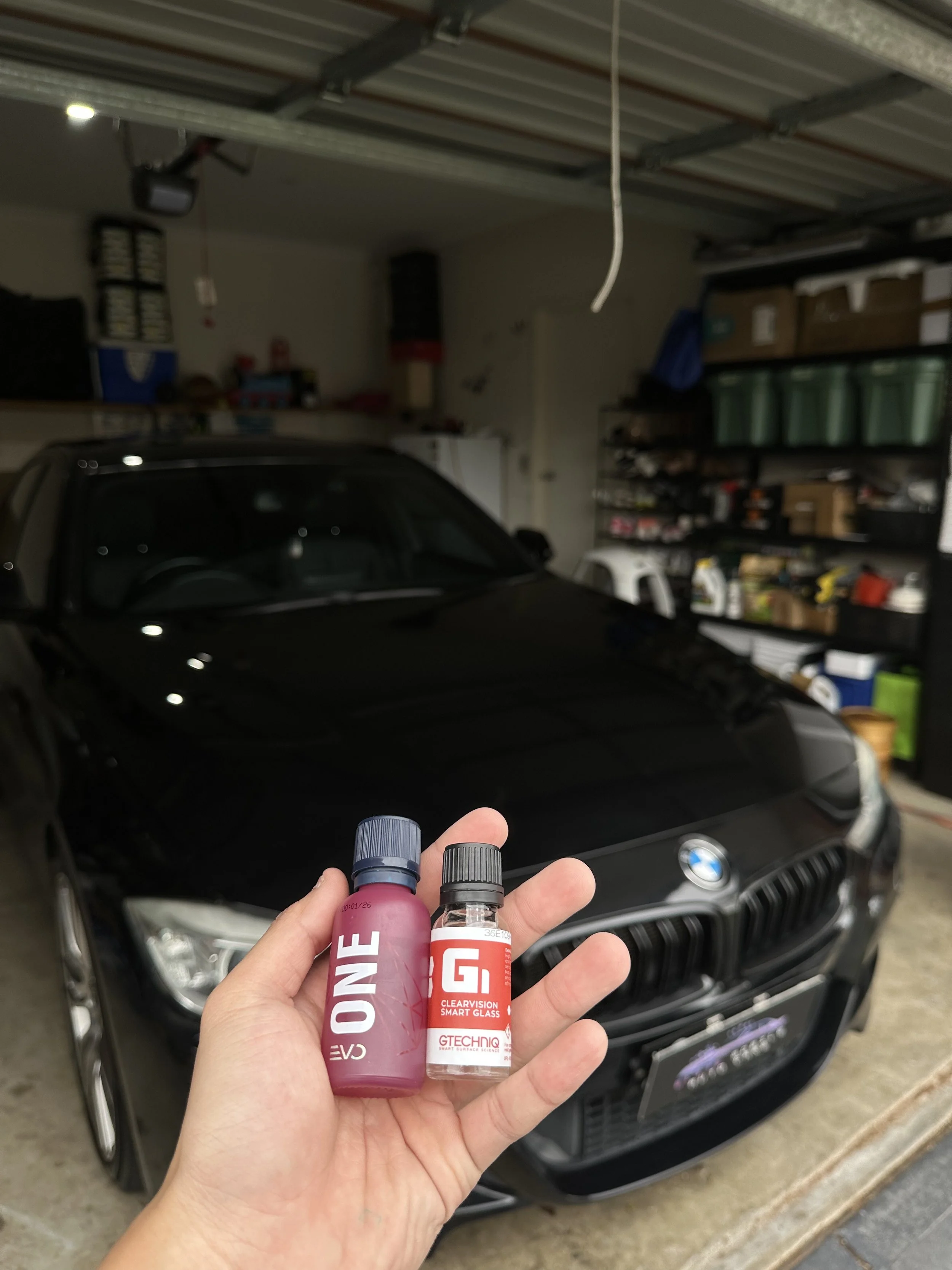 Hand holding two bottles of glass cleaner in front of a black BMW car parked in a garage with storage shelves.