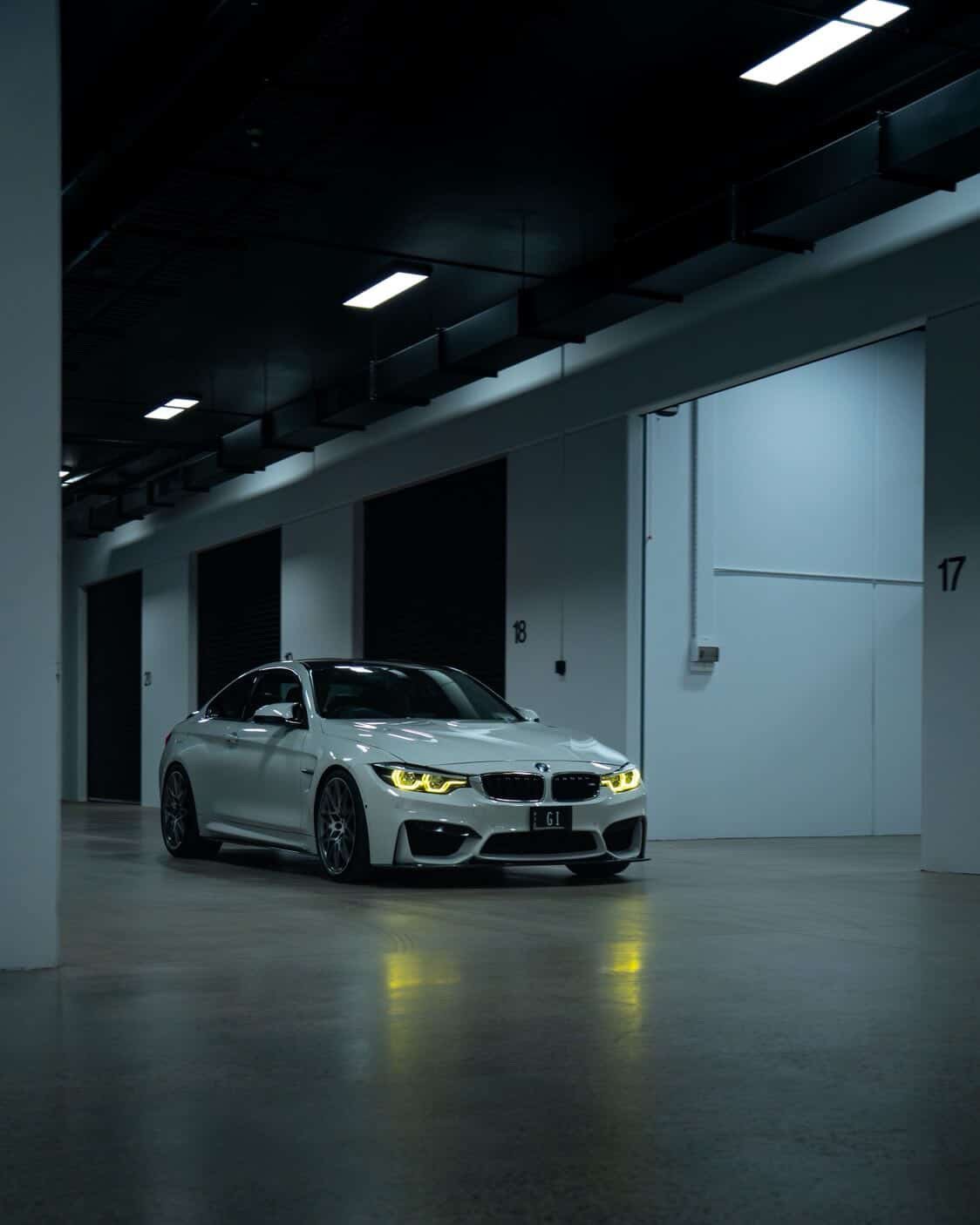 A white BMW car parked inside a dimly lit indoor parking garage with concrete flooring and white walls, the car just got a full exterior detail by Burwood car detailer
