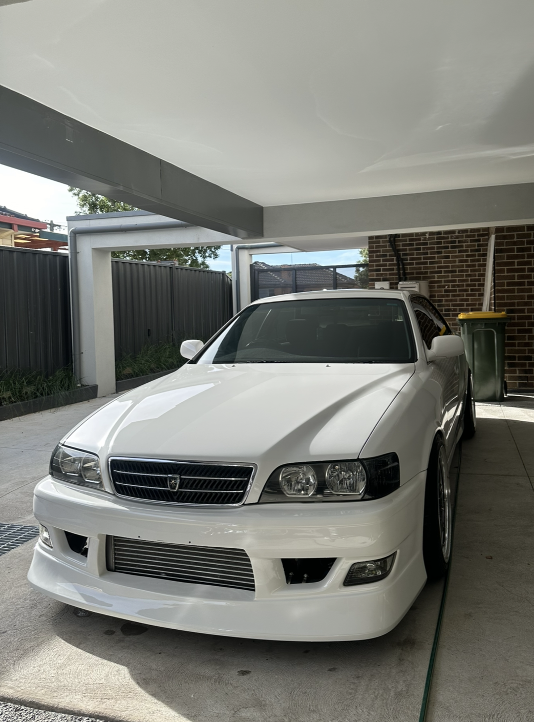 White car parked under a carport with brick wall and yellow trash bin in background.