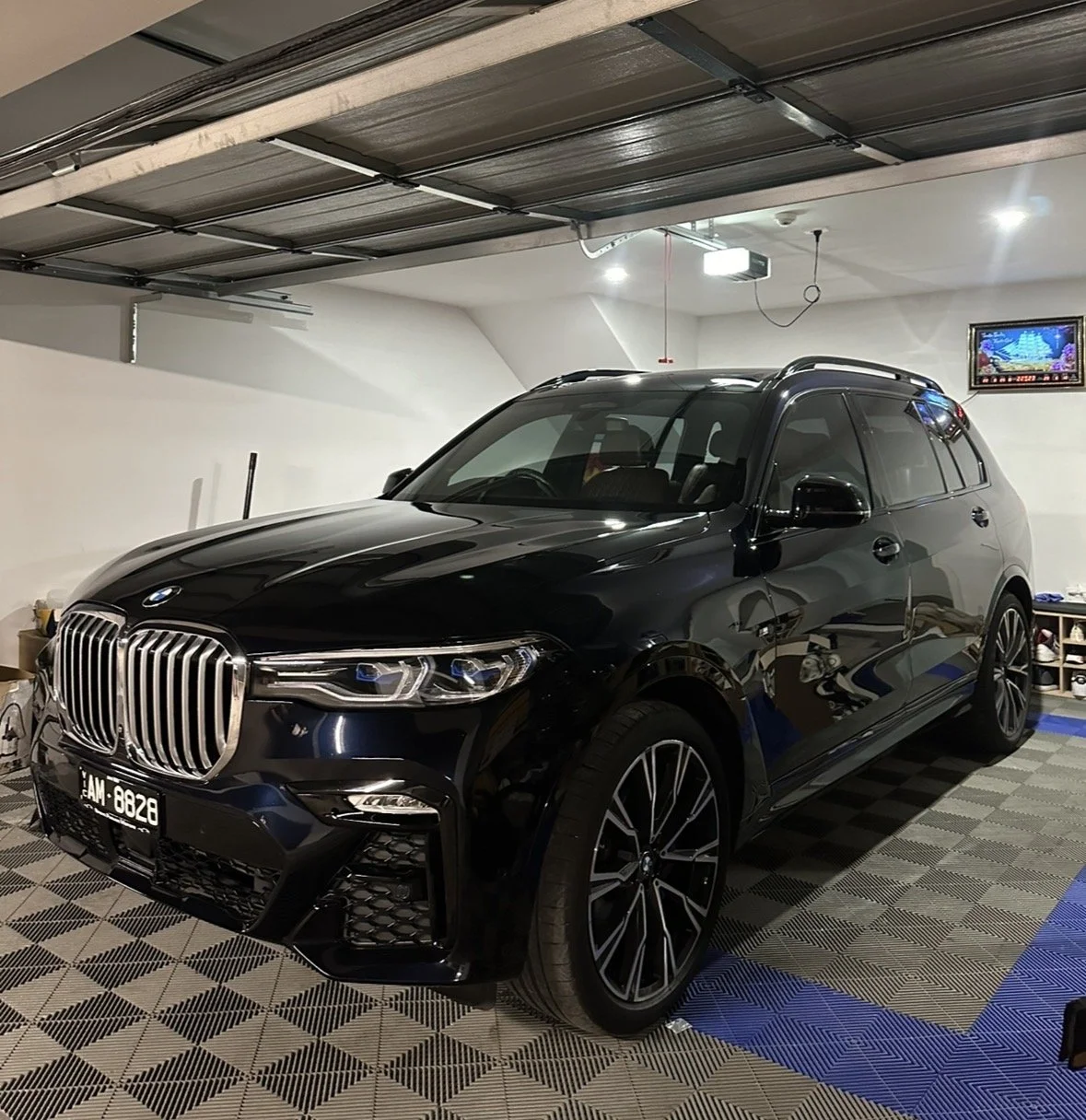 Black luxury SUV parked in a garage with white walls, fluorescent lighting, a small TV on the wall, and items on a shelf in the background.