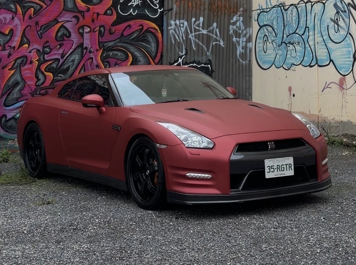 A red Nissan GT-R sports car parked on gravel in front of a graffiti-covered wall.