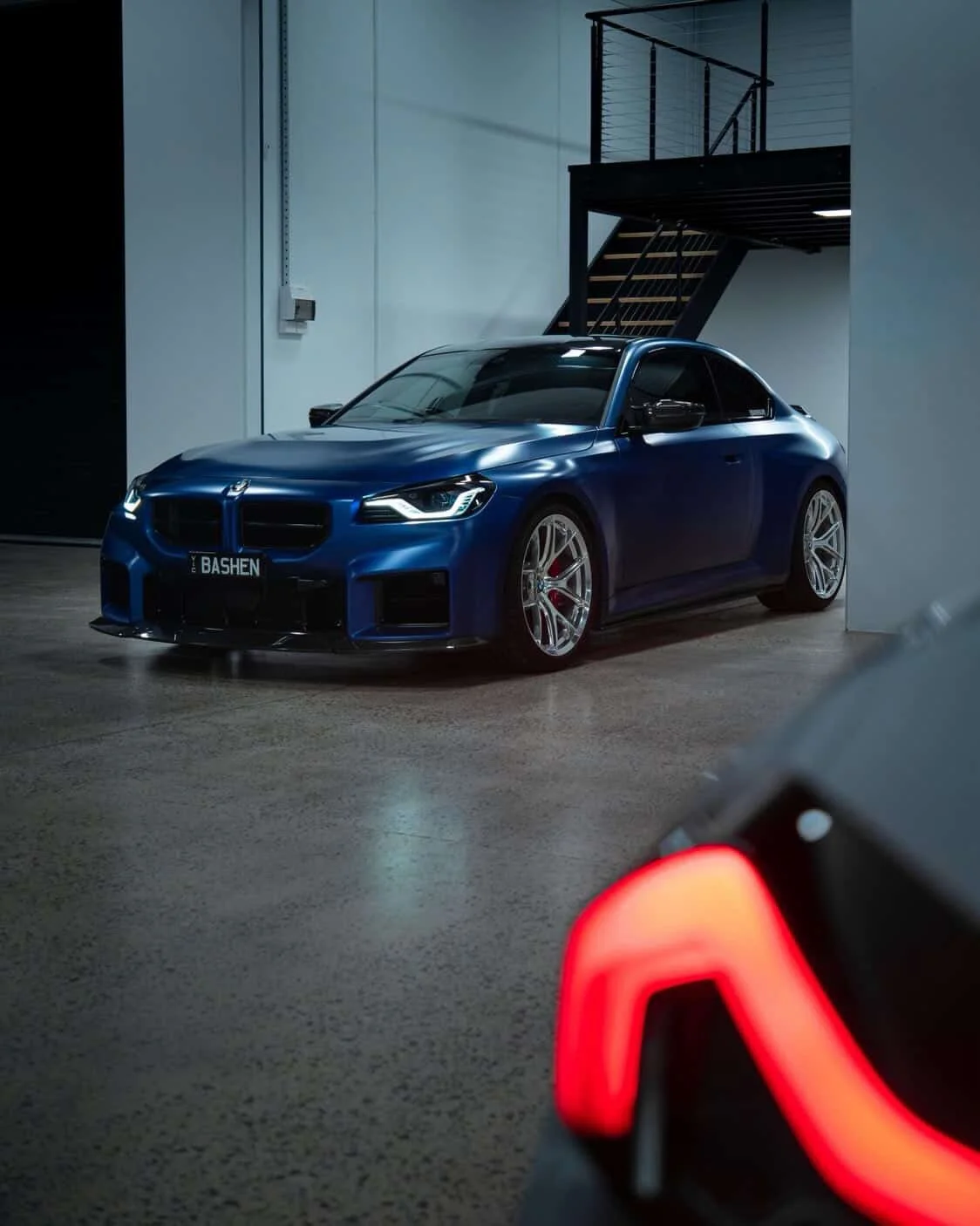 A blue sports car with a license plate reading 'BASHEN' parked indoors near a staircase, with a partial view of another vehicle's rear light in the foreground.