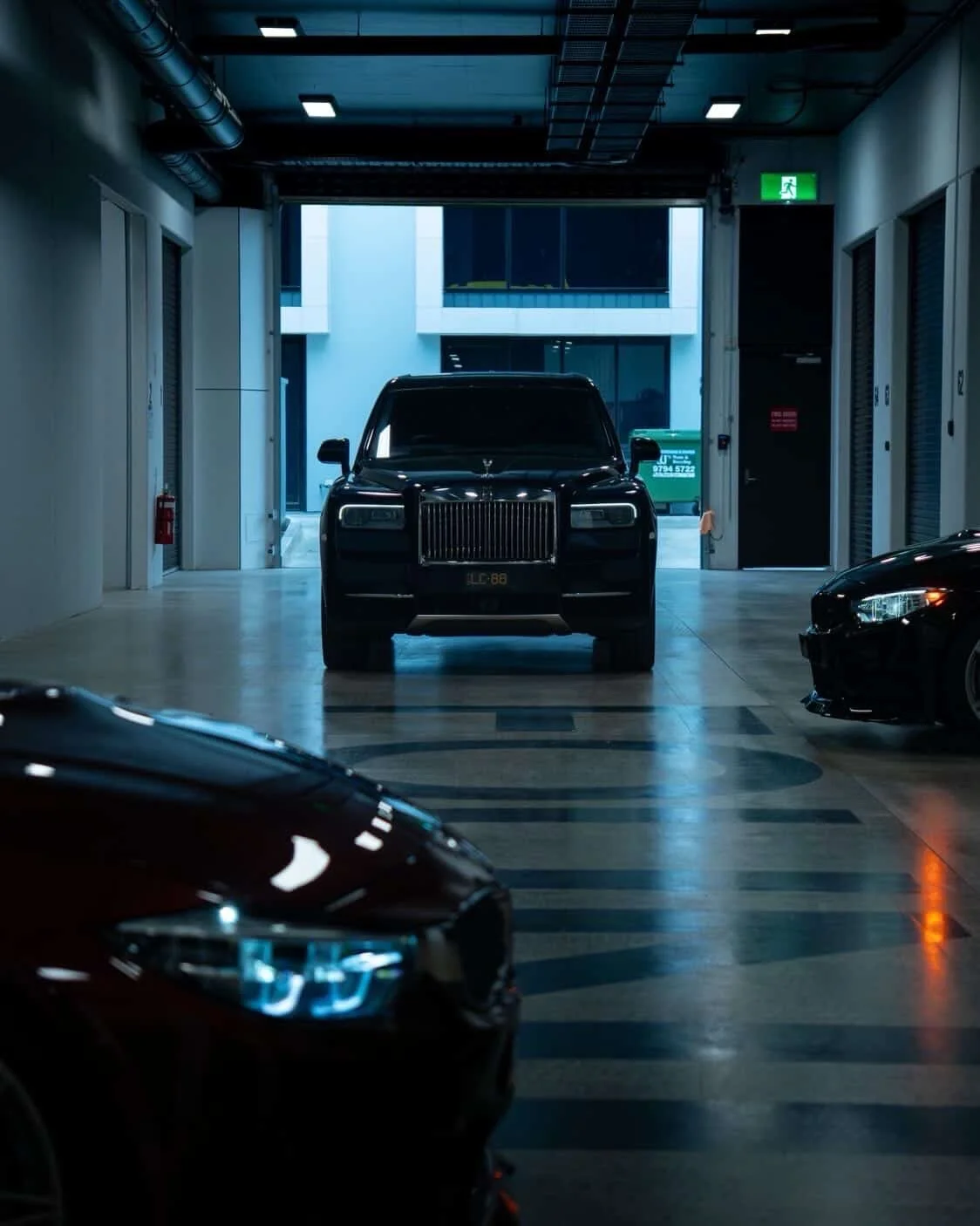A black luxury SUV parked inside a modern indoor parking garage with a high ceiling, visible pipes, and an open garage door showing buildings outside. Part of another car is visible in the foreground. All cars were ceramic coated by mobile detailer