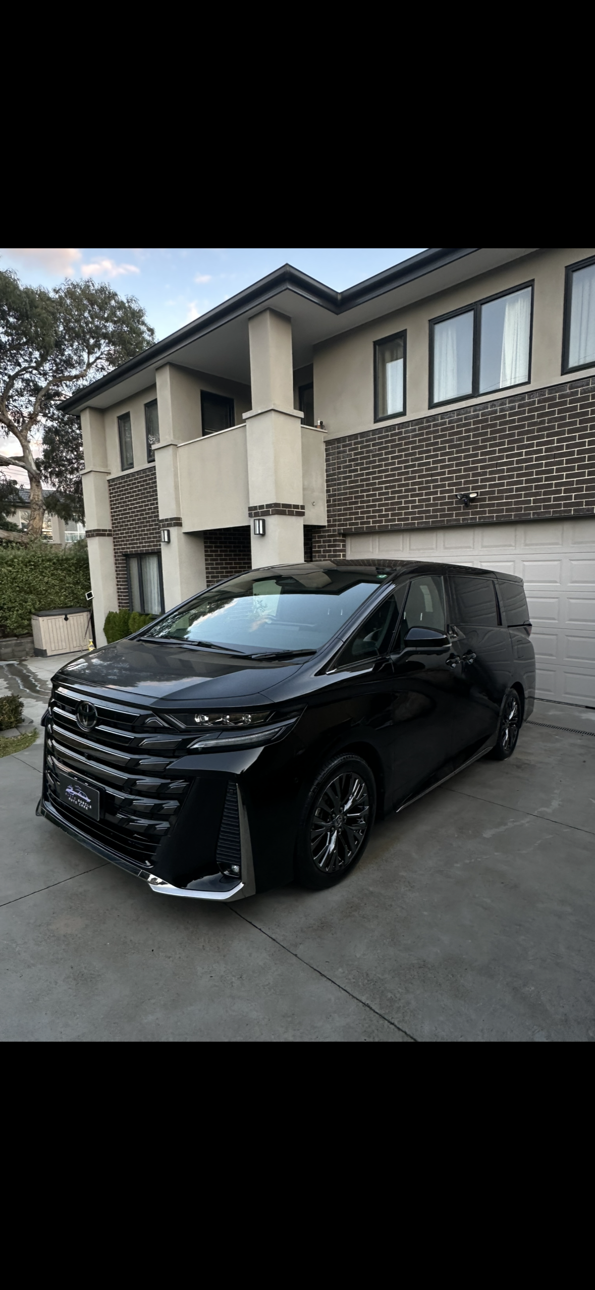 Black Toyota minivan parked in driveway in front of a modern, two-story house with brick and stucco exterior, multiple windows, and a white garage door.