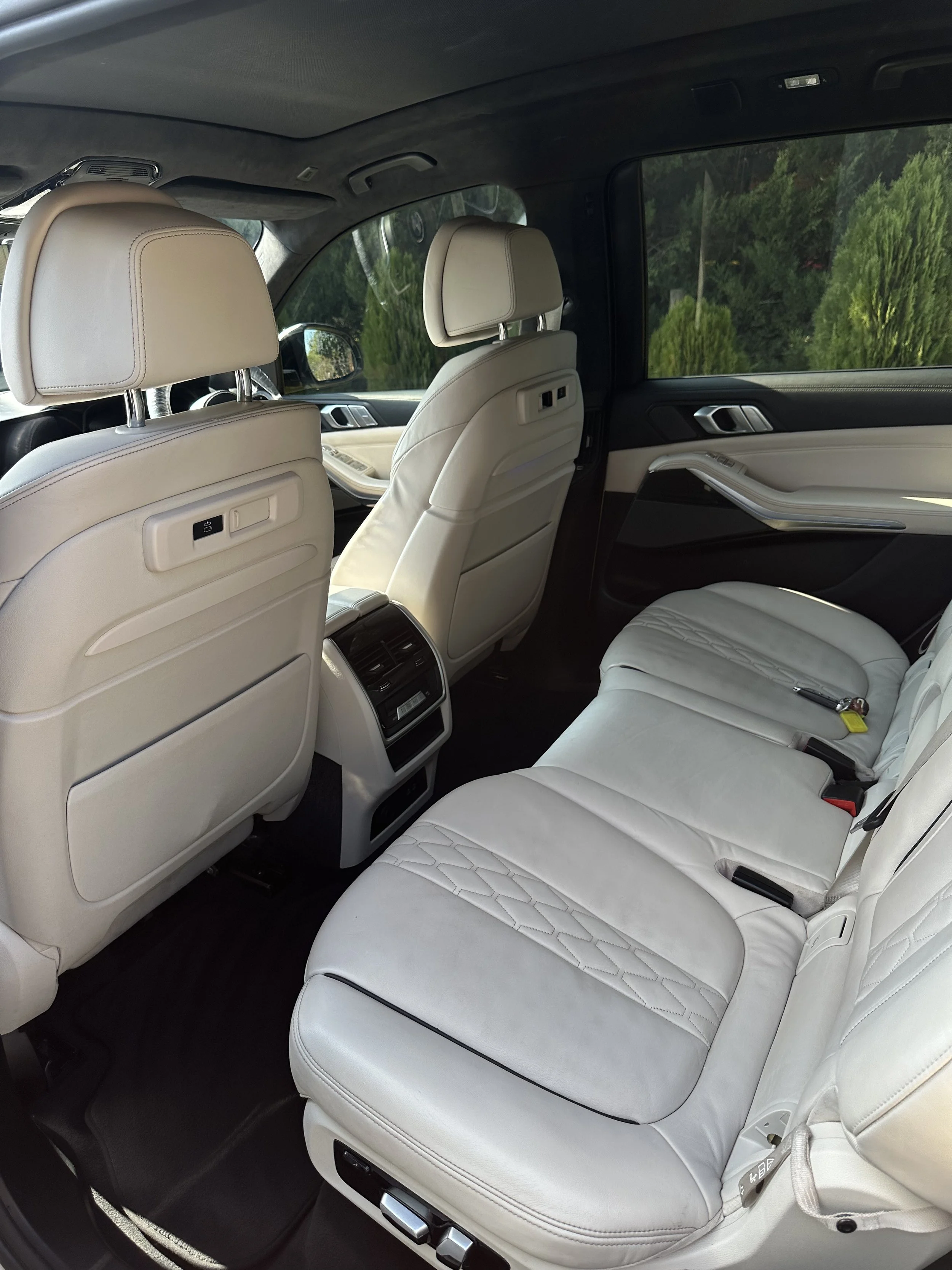 Interior of a luxury car with white leather seats, black dashboard, and green trees visible outside the windows.