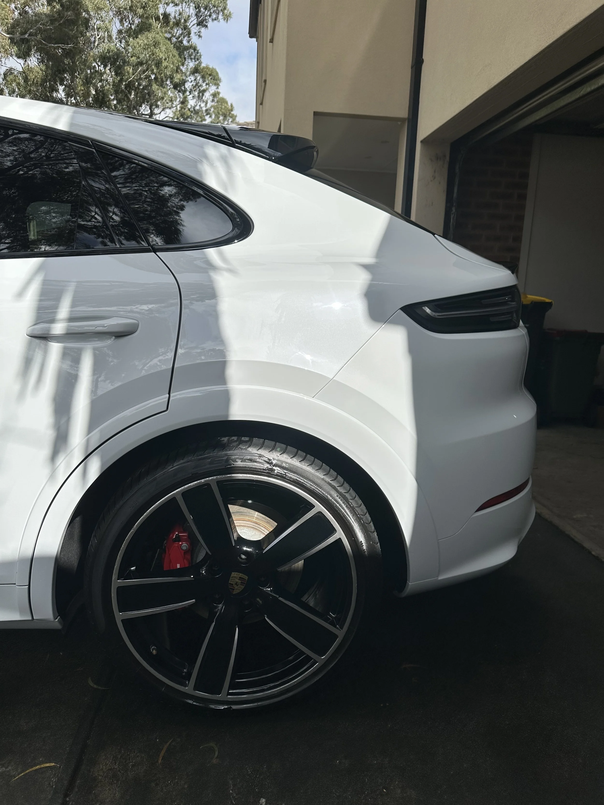 Close-up side view of a white luxury car parked in a driveway, showing a black wheel with red brake caliper, part of the rear window, and a garage in the background.