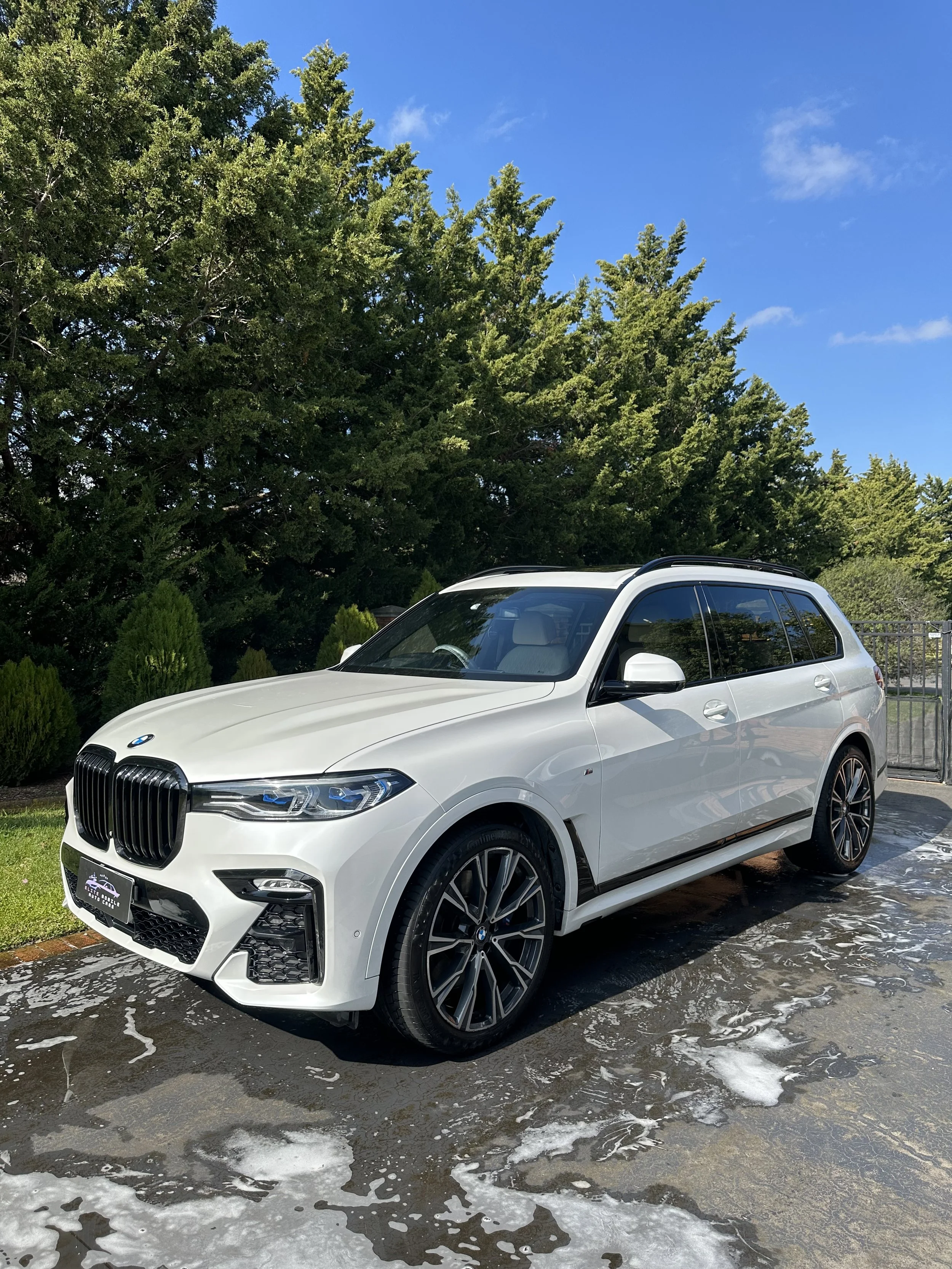A white BMW SUV is parked outside on a wet pavement with soap suds, under a clear blue sky with a few clouds, surrounded by green trees and shrubbery.