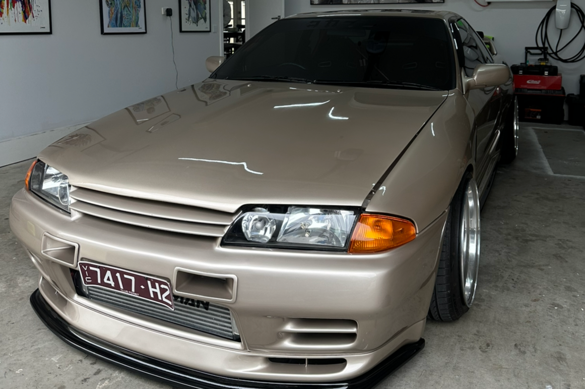 A beige modified Nissan sports car with an aftermarket front bumper, low profile tires, and custom wheels parked inside a garage with a paint protection coating
