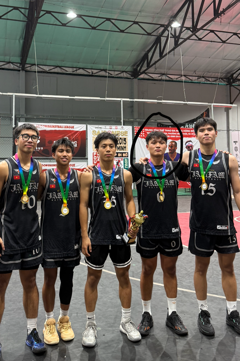 Group of five young male basketball players in black uniforms with medals around their necks, one holding a trophy, standing on a basketball court inside a gymnasium.