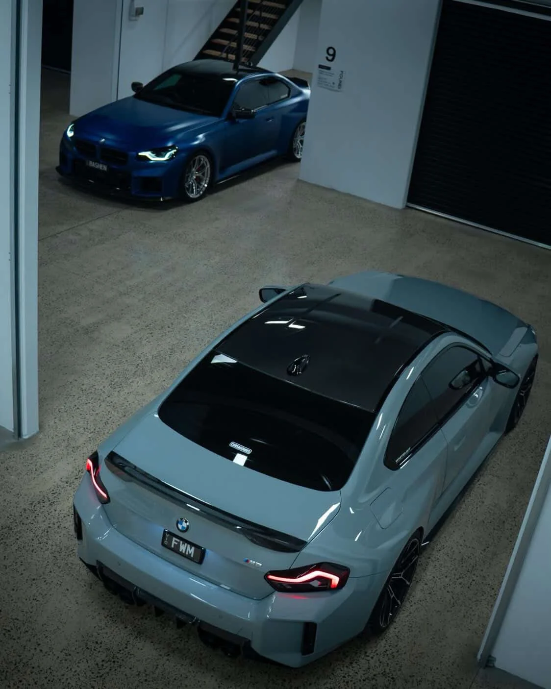 Two BMW cars parked indoors, one in the foreground with a silver body and black roof, the other in the background with a blue body, near a staircase and a wall with an elevator sign.