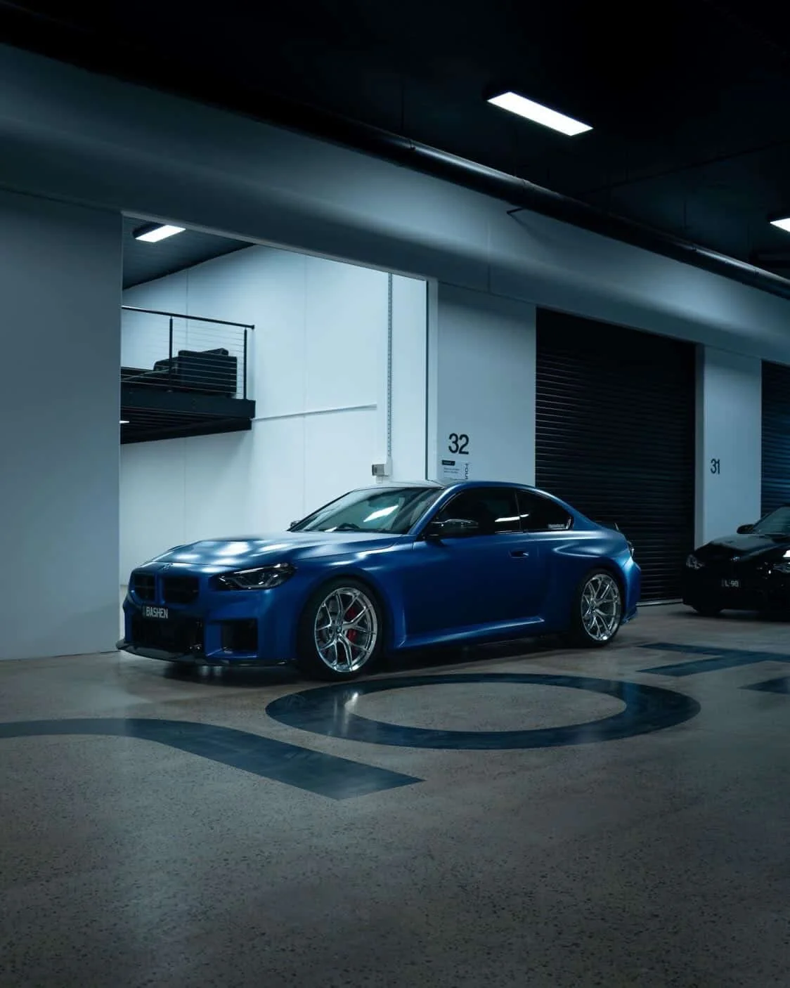 A blue sports car parked in an indoor parking garage with illuminated ceiling lights and numbered parking spots 32 and 31.