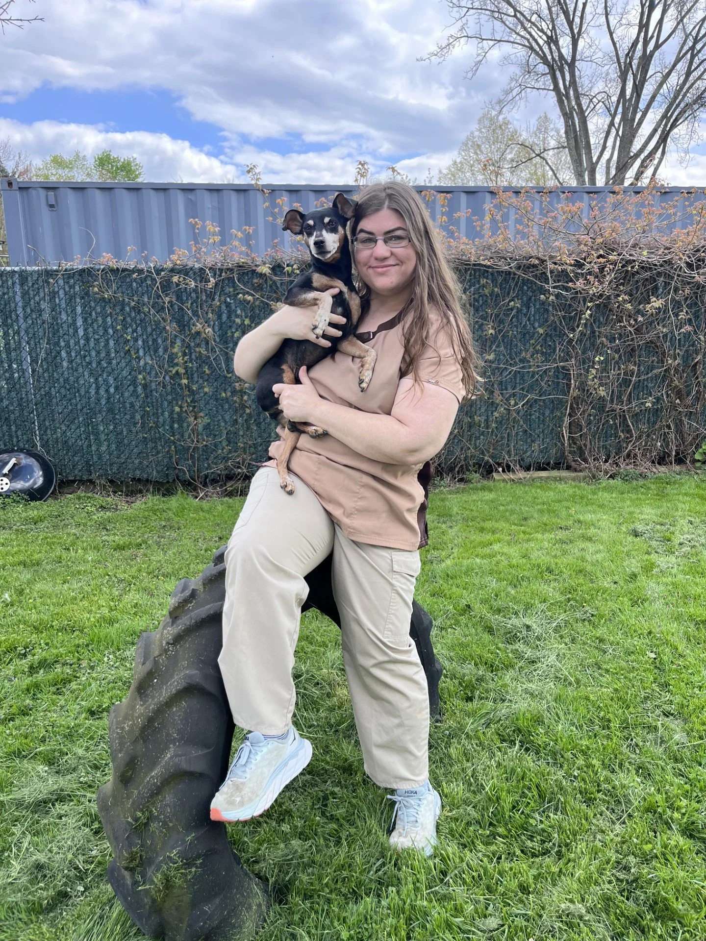 A woman holding a small black and tan dog while sitting on a large tractor tire in a grassy backyard with a chain-link fence and leafless trees under a cloudy sky.