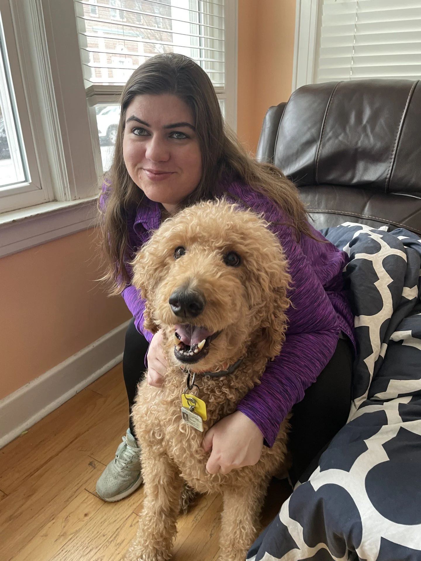 A woman with long brown hair and blue eyes is sitting on a brown leather couch next to a window with white blinds, holding a smiling curly-haired beige dog with a yellow collar and a pet tag.