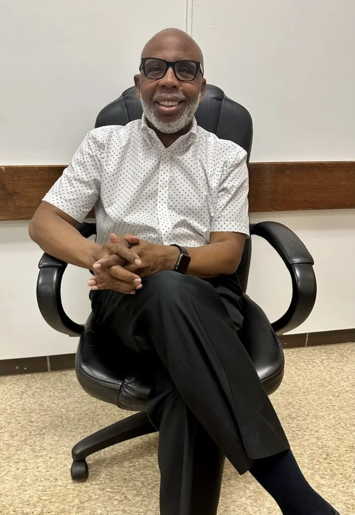 Smiling man with gray beard and glasses sitting in an office chair, wearing a white polka dot shirt and black pants, in front of a white wall with a wooden trim.