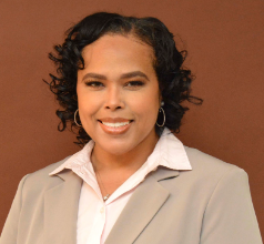 Portrait of a woman with curly dark hair wearing a beige blazer and white collared shirt, smiling against a brown background.