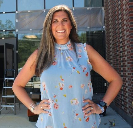 A woman with long brown hair wearing a sleeveless blue floral top, standing outdoors with a brick building and glass structure in the background, smiling and posing with hands on hips.