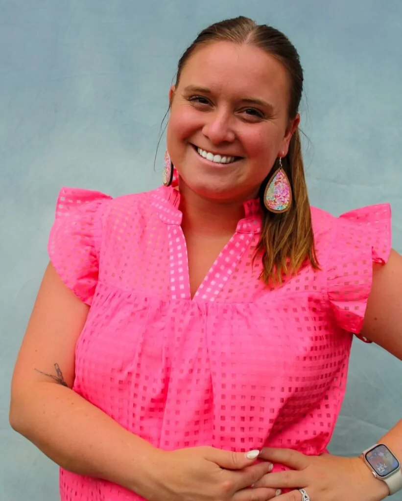 Smiling woman with brown hair wearing pink dress with ruffled sleeves, large colorful earrings, and a smartwatch, standing against a light blue background.