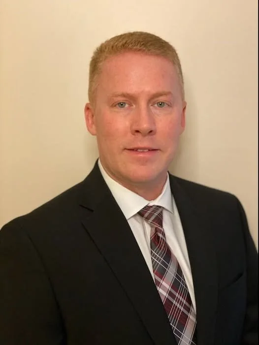 A portrait of a man with short blond hair wearing a dark suit, white dress shirt, and a plaid tie, standing against a plain light-colored wall.