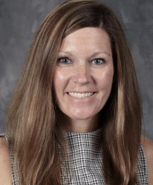 A woman with long, brown hair smiling at the camera, wearing a checkered sleeveless top against a gray background.