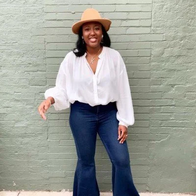 A woman wearing a wide-brimmed beige hat, white blouse, and dark blue flared jeans standing in front of a light green brick wall, smiling.