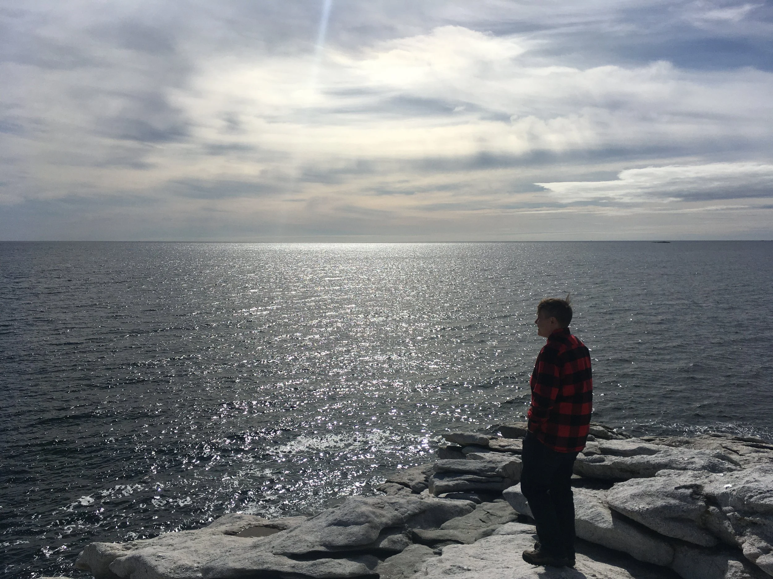 Adrienne, wearing a red and black checkered flannel shirt, stands on rocks by the ocean, looking out over the water under cloudy skies.