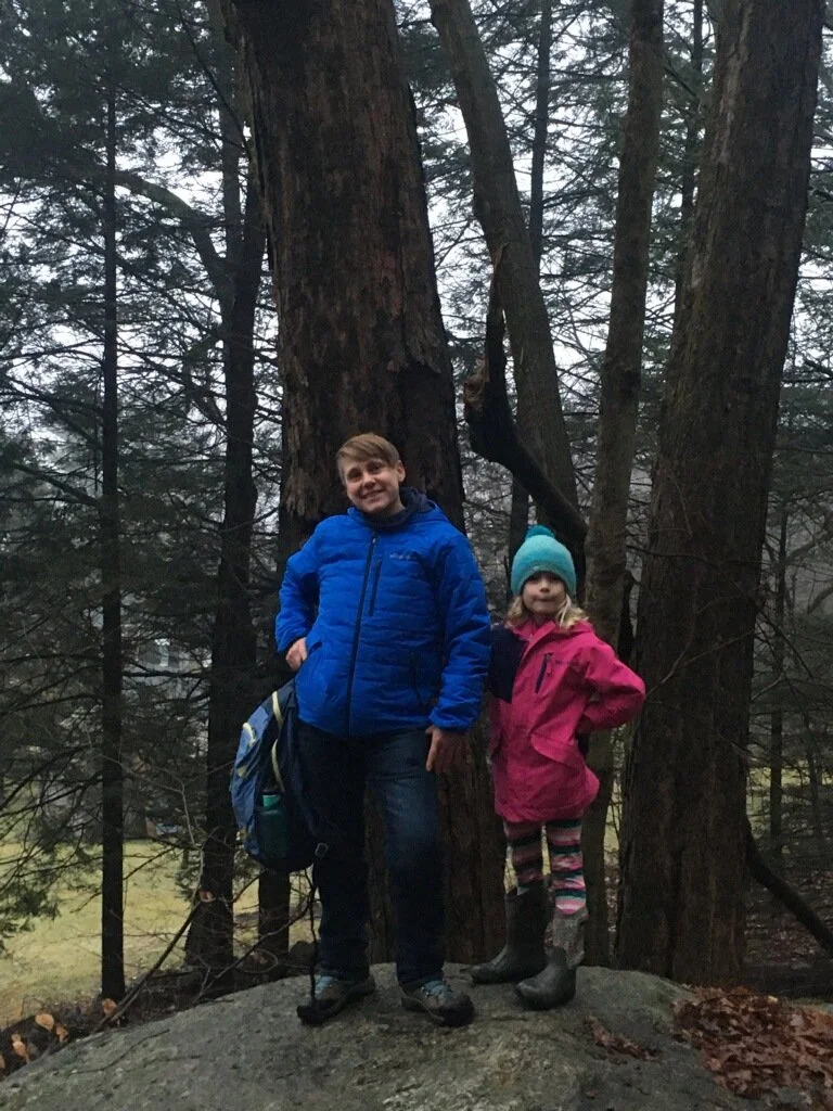 Adrienne and their daughter standing together on a rock in a forest with tall trees and misty background.