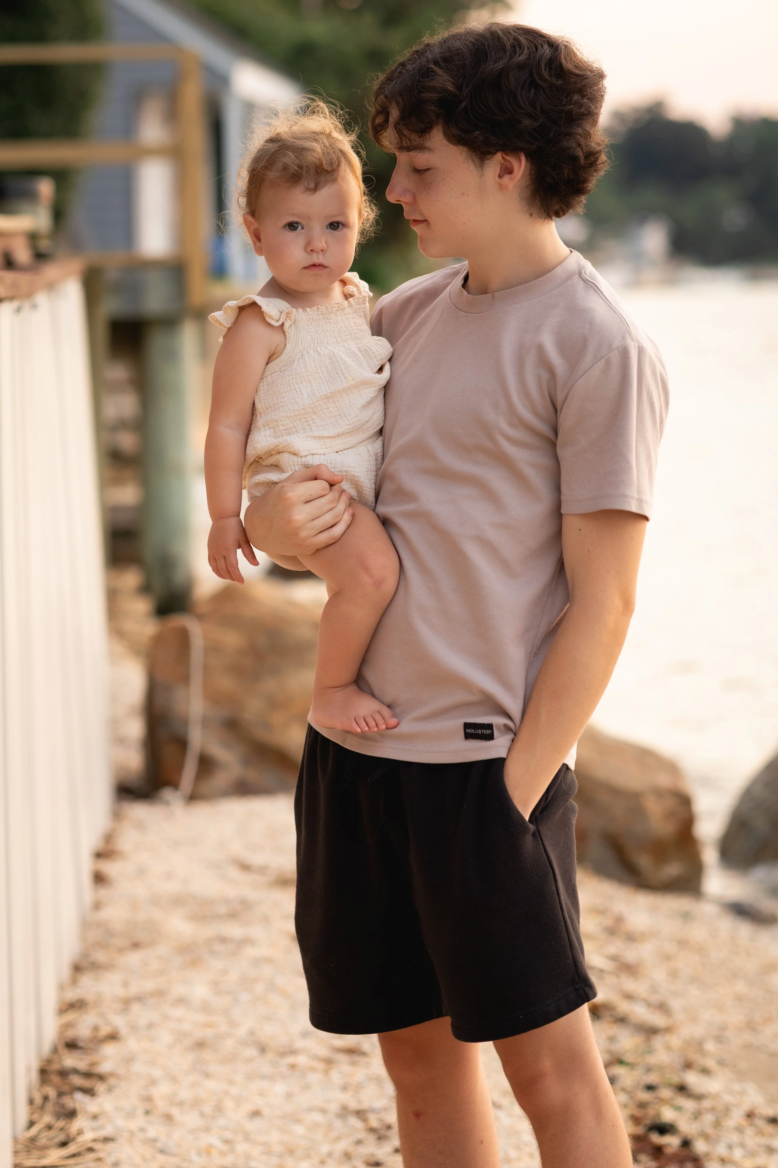 A young man holding a small girl in his arms on a beach, with rocks and water in the background, during sunset.