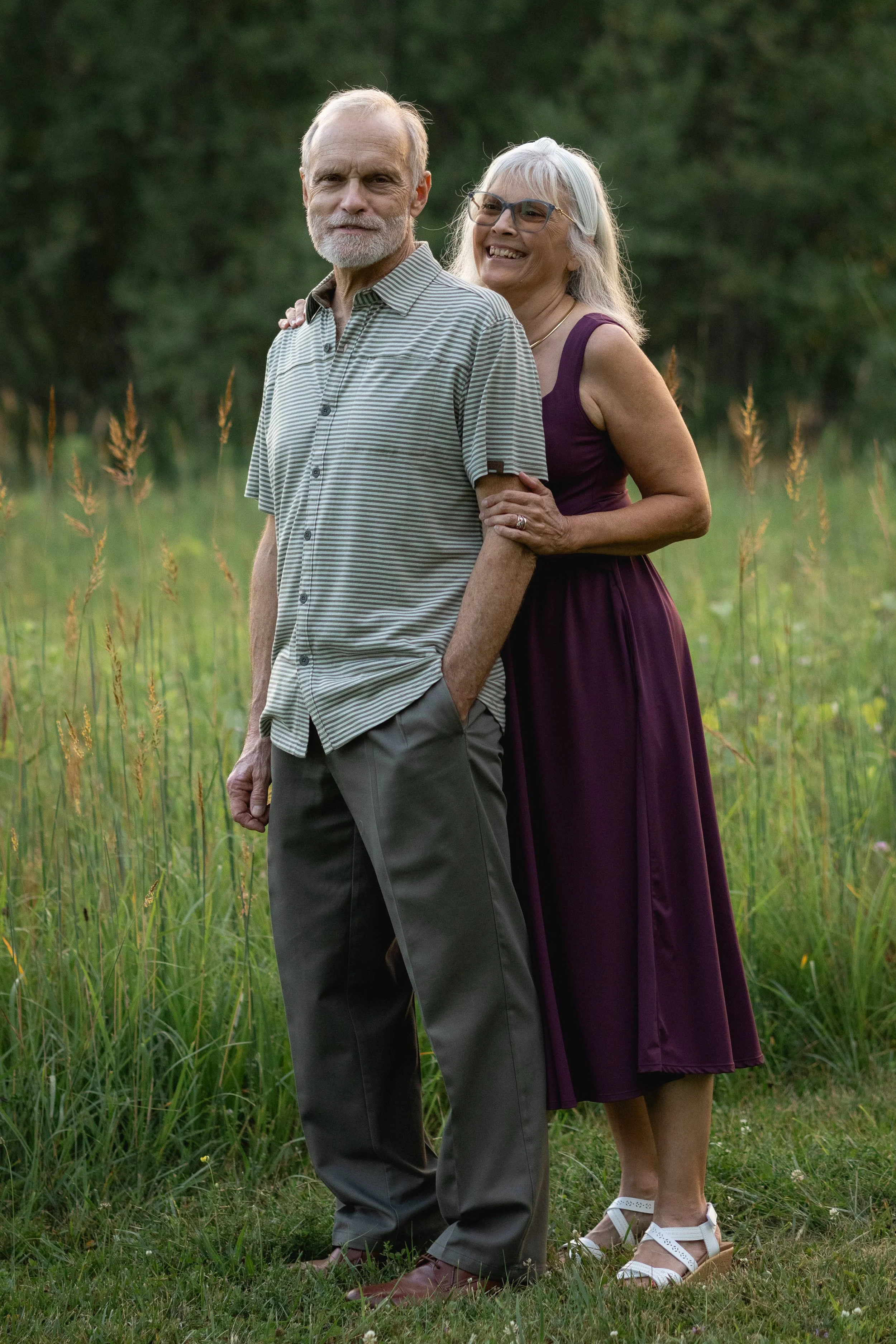 A happy elderly couple stands together outdoors in a grassy field surrounded by trees during daytime. The woman is smiling, holding onto the man's arm, and wearing glasses and a purple dress. The man has a beard, wears a striped short-sleeved shirt and gray pants, and has his hands in his pockets.