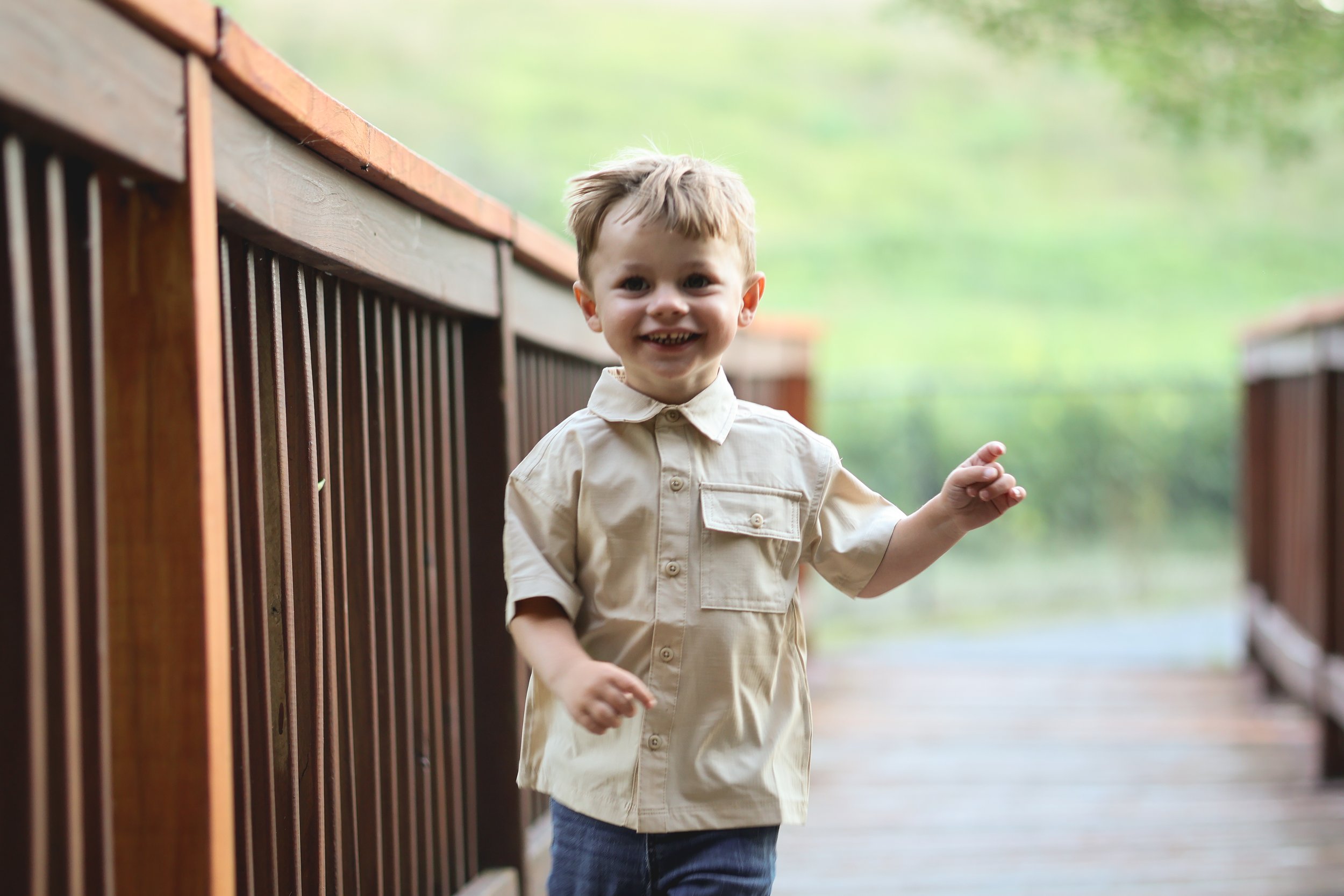 A young boy with light brown hair and a big smile walking along a wooden bridge outdoors, wearing a beige button-up shirt and blue jeans, with green trees and a blurred background.