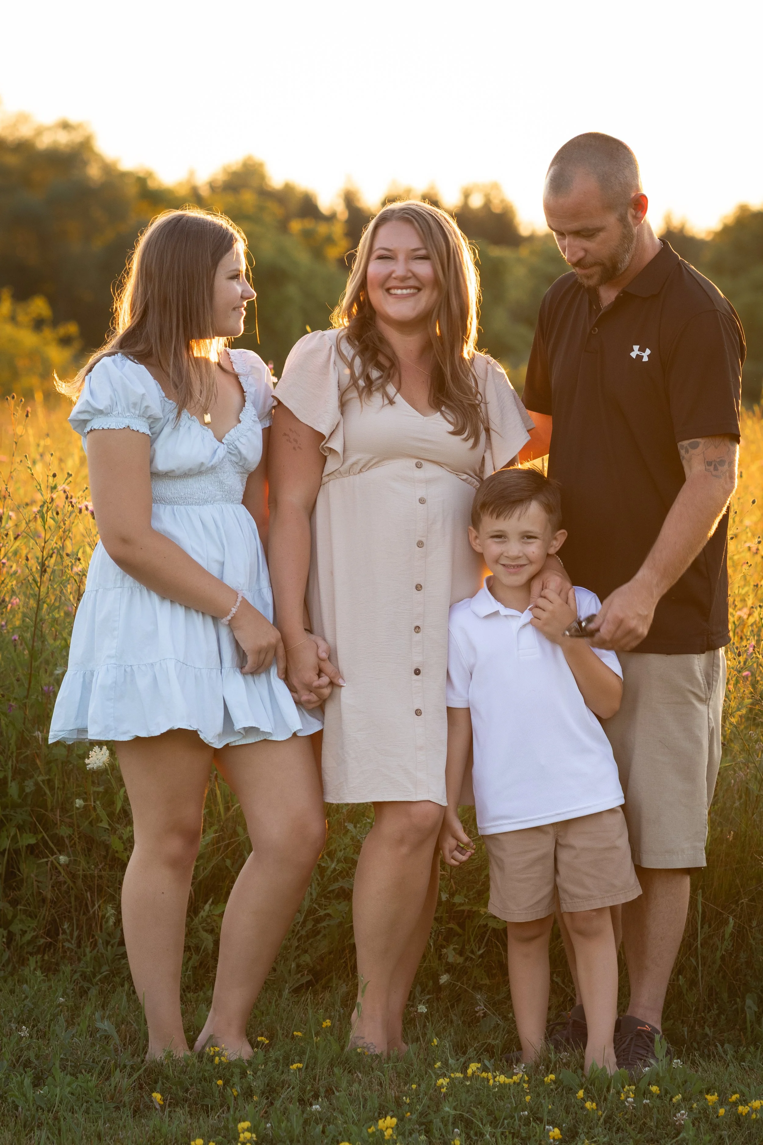 A family of four standing in a grassy field with yellow flowers, during sunset, smiling and looking at each other.