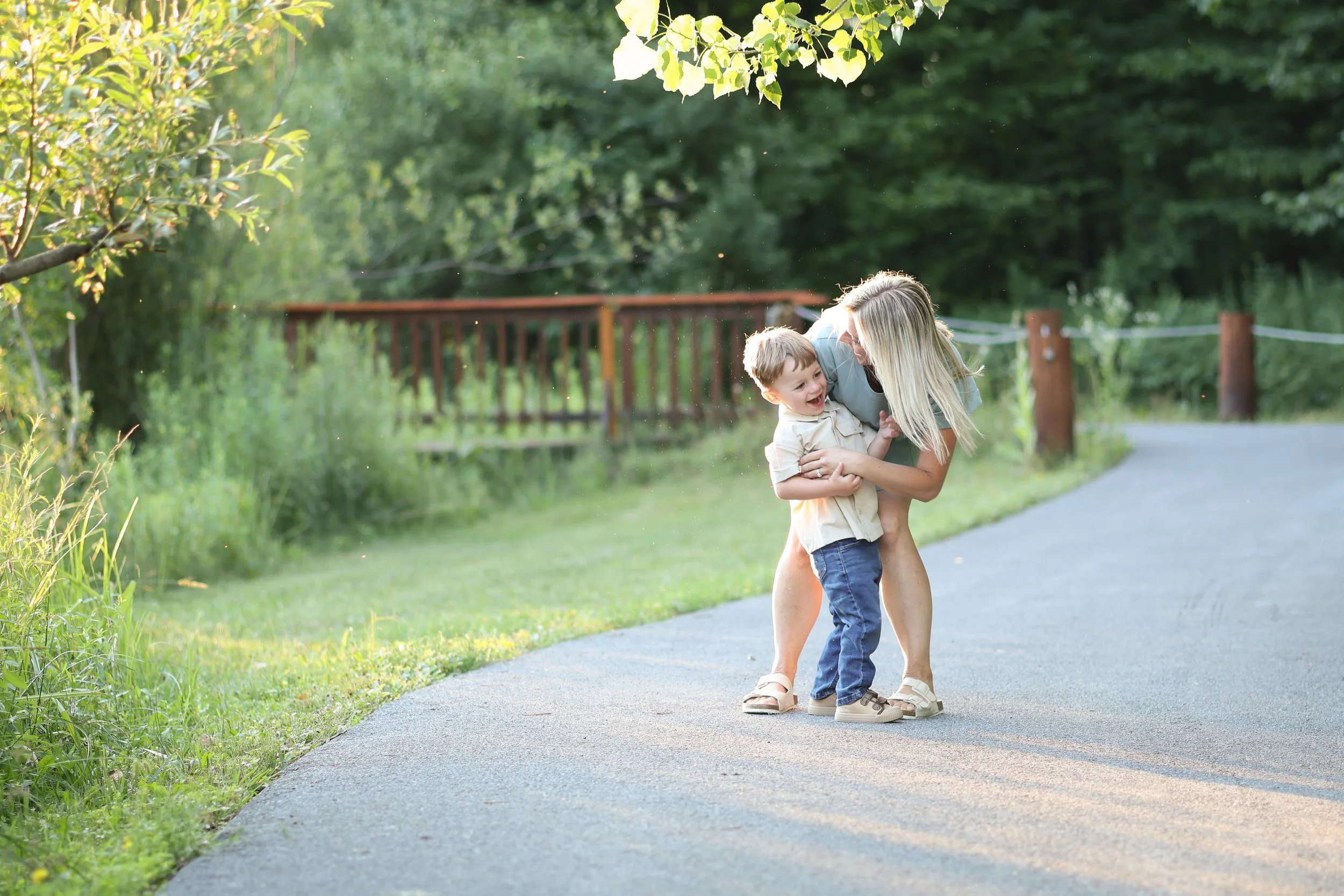 A woman and young boy are joyfully playing and laughing outdoors on a paved path surrounded by green trees and grass, with a wooden fence in the background.