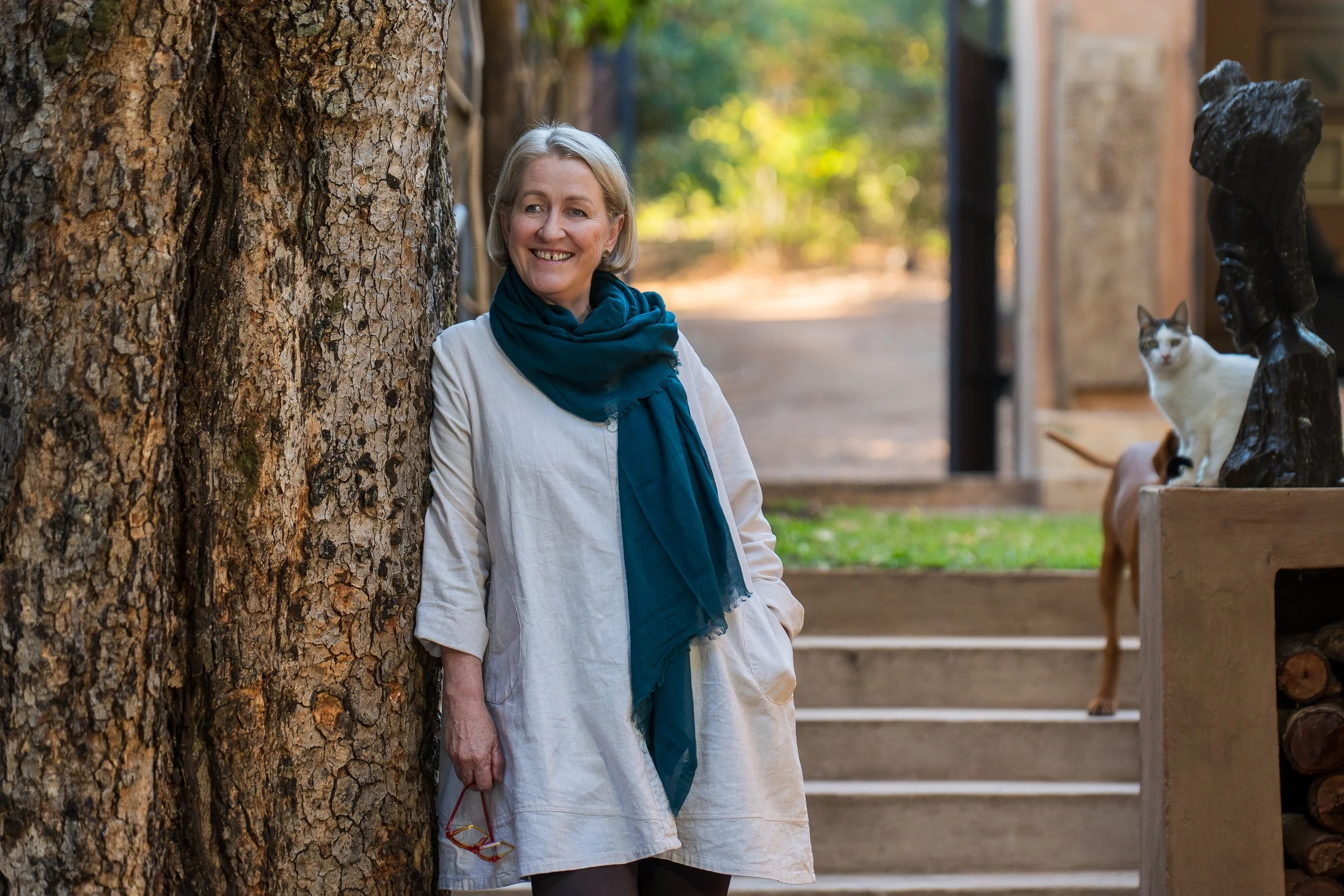 A woman with gray hair in a white jacket and teal scarf smiling and leaning against a tree in an outdoor setting with a cat and a dog nearby.