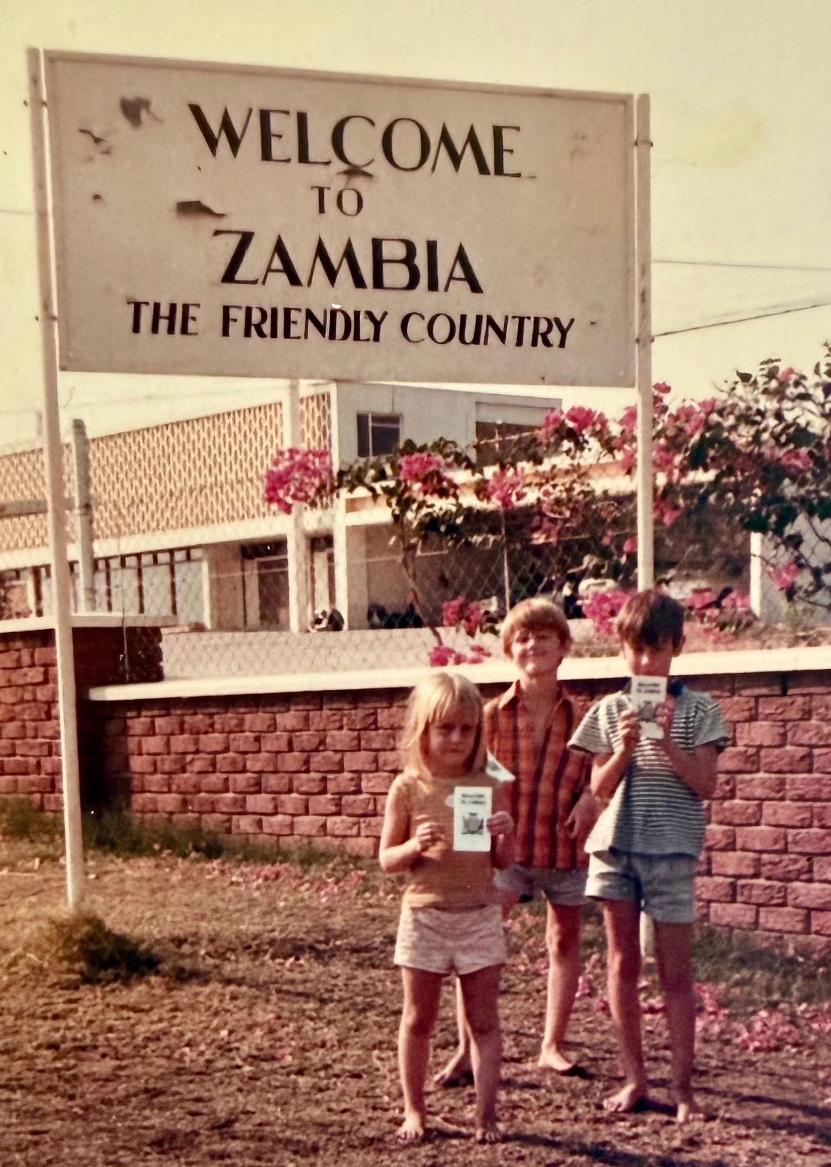 Three children standing outside in front of a brick wall and a welcome sign that reads 'Welcome to Zambia the Friendly Country,' holding papers or photographs.